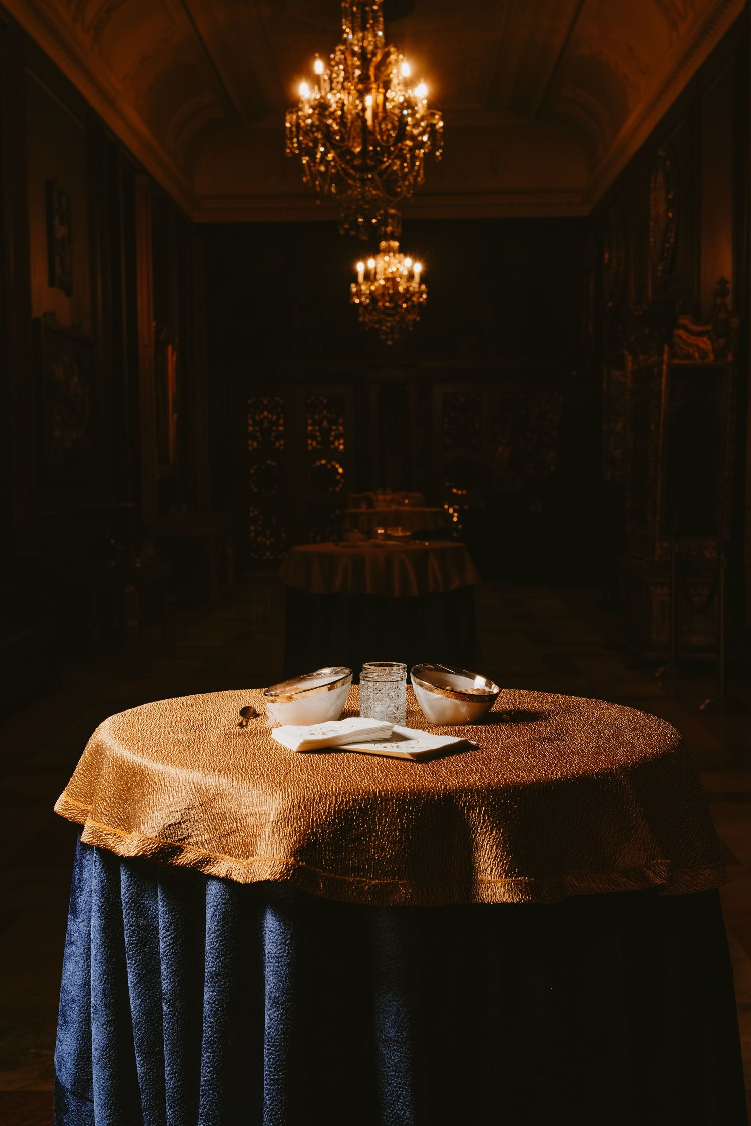 Elegant dining tables with blue tablecloths and gold overlays illuminated by crystal chandeliers in a dimly lit room.