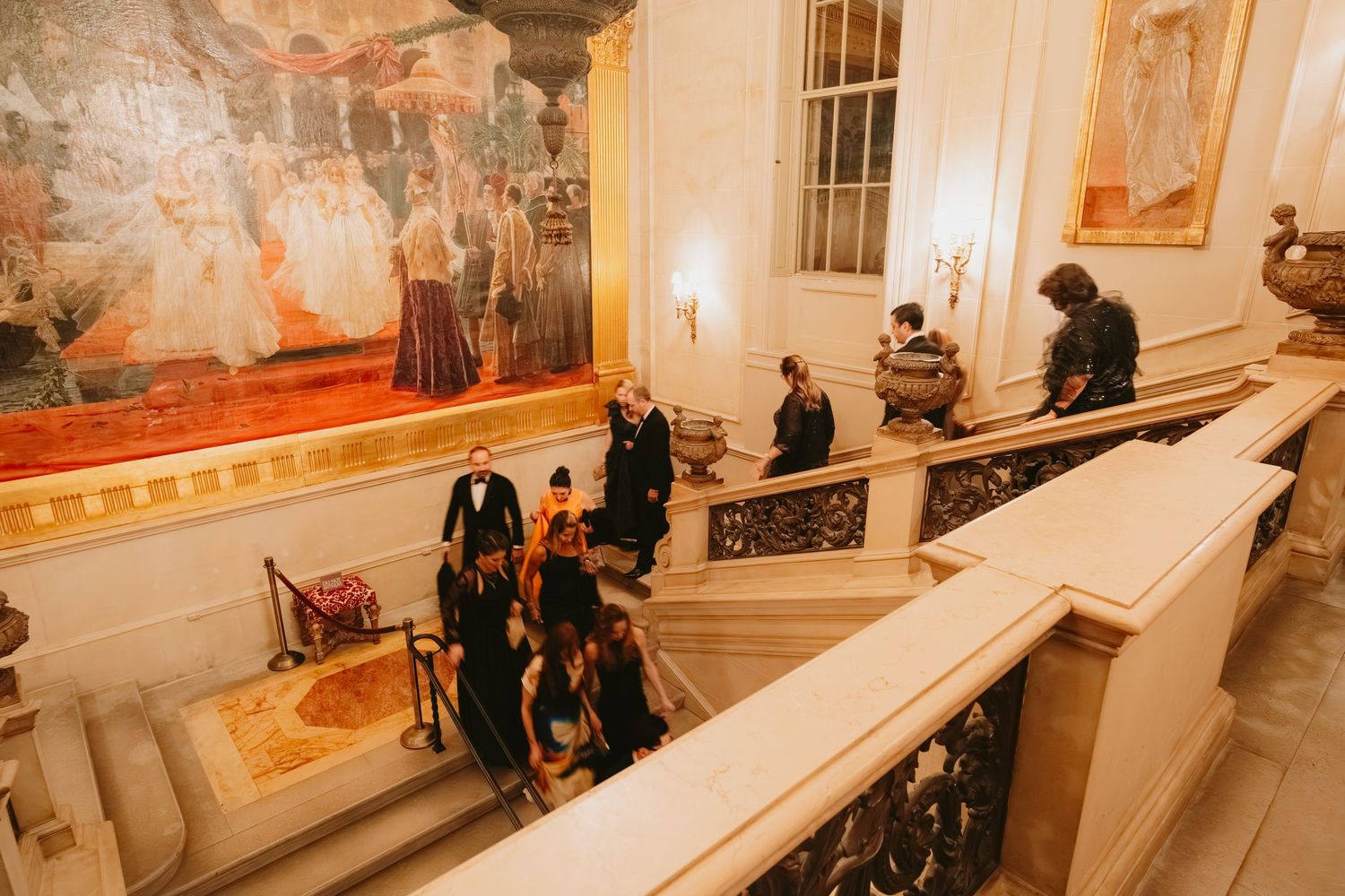 People gather on an elegant marble staircase inside a historic building with ornate murals and decorative details.