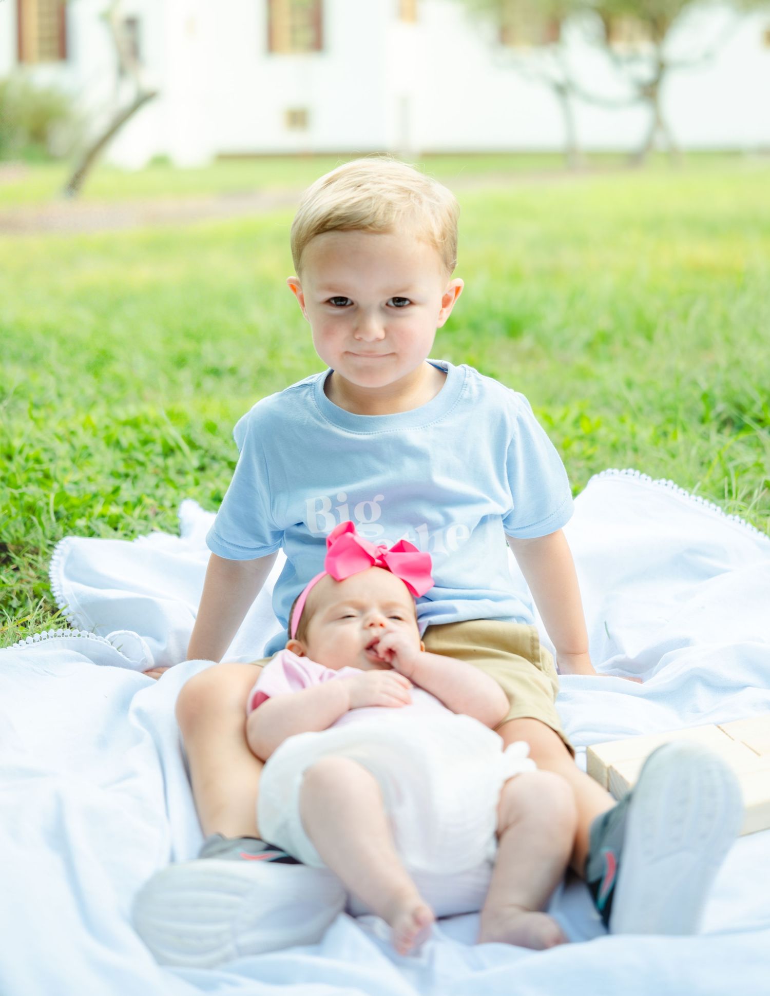Two children sit together on a white blanket laid out on grass on a sunny day.