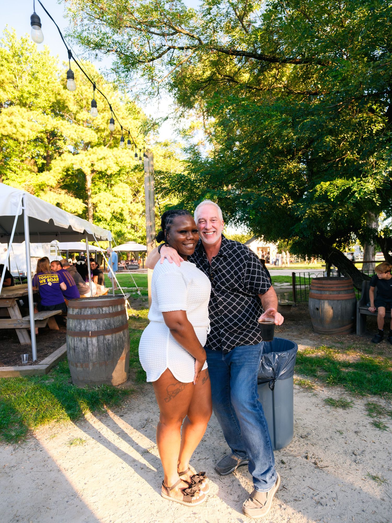 Friends gathered near wooden barrels at an outdoor summer event with trees providing shade.