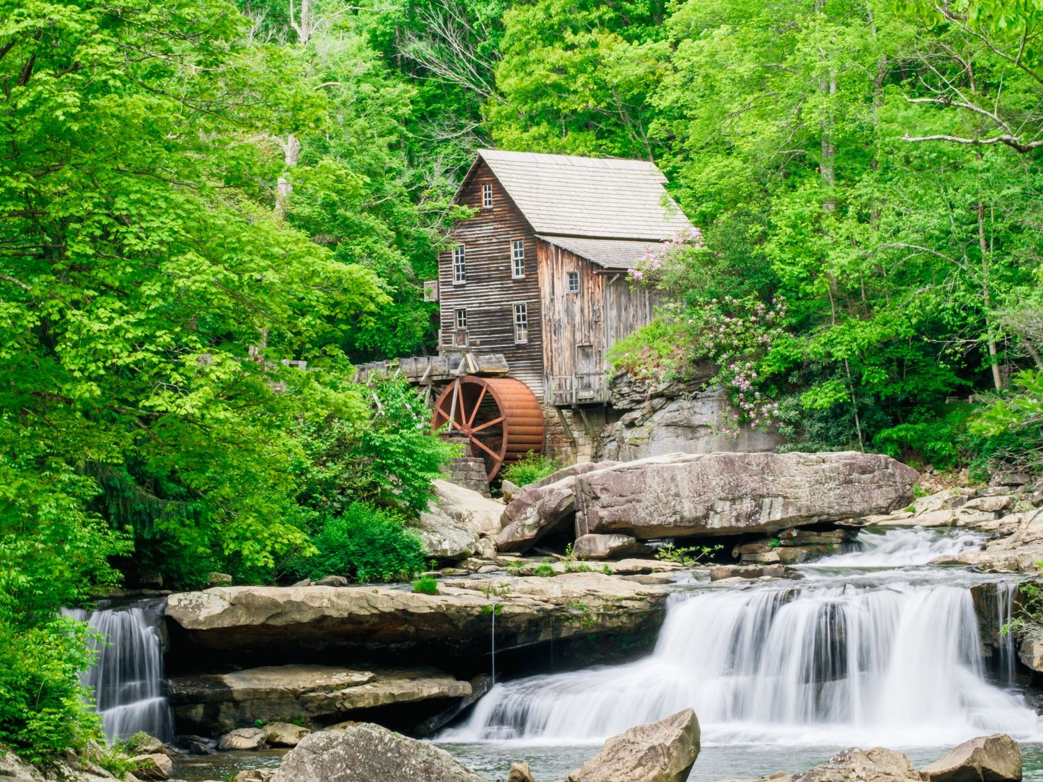 Old rustic grist mill with wooden water wheel perches above cascading waterfalls surrounded by lush green forest.
