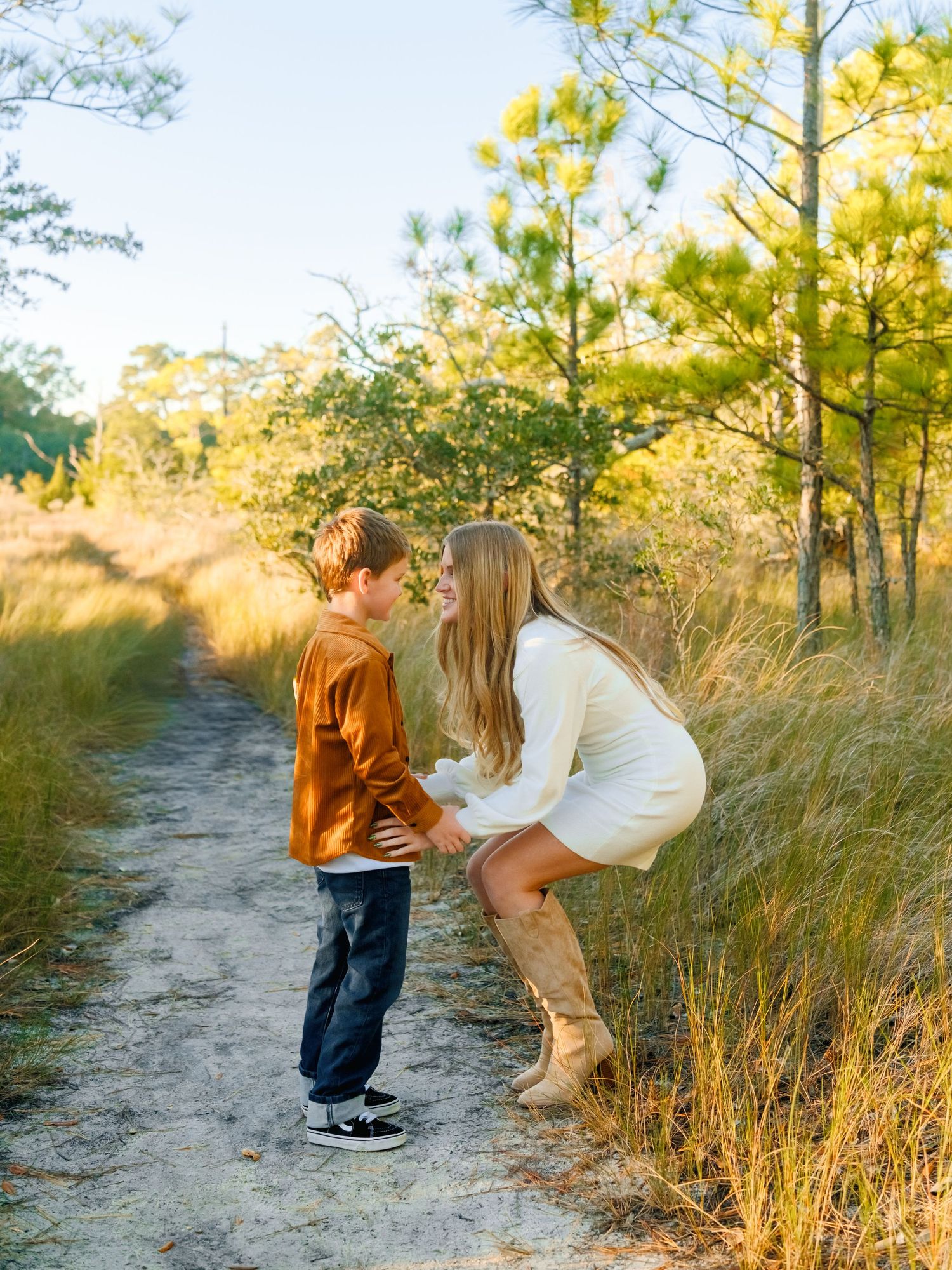 A mother and child share a tender moment on a sandy path surrounded by tall grass and pine trees at sunset.