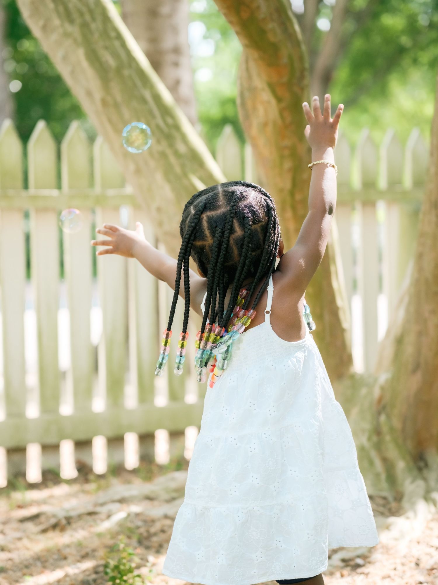 A child in a white dress runs and plays with bubbles in a backyard with white picket fence and green trees.
