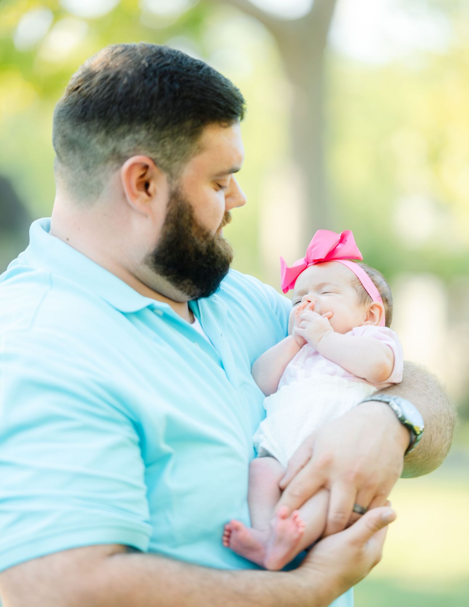 A bearded man in a turquoise polo shirt tenderly holds a sleeping baby wearing a pink bow headband outdoors.