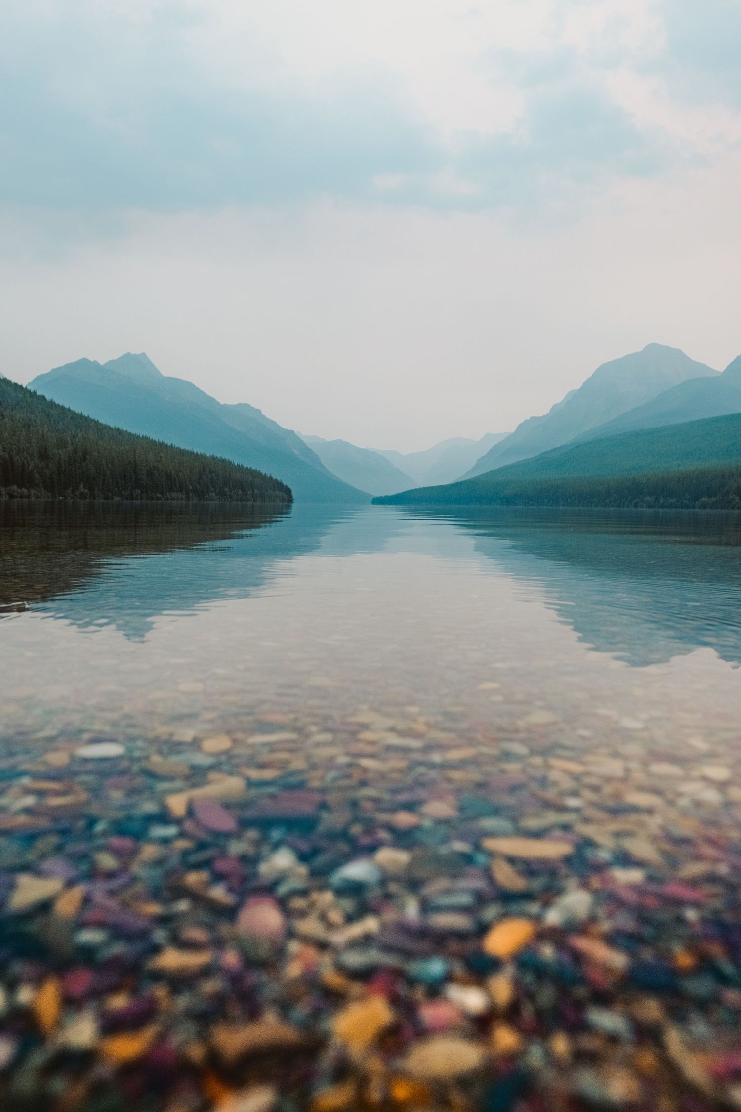 Split-focus landscape photo showing colorful pebbles underwater and misty mountains rising above a tranquil lake.