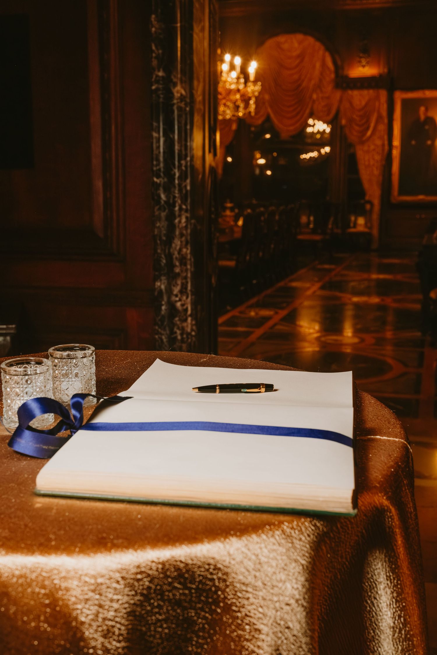 A wedding guest book and pen rest on an elegant table with a white tablecloth in a dimly lit reception hall.