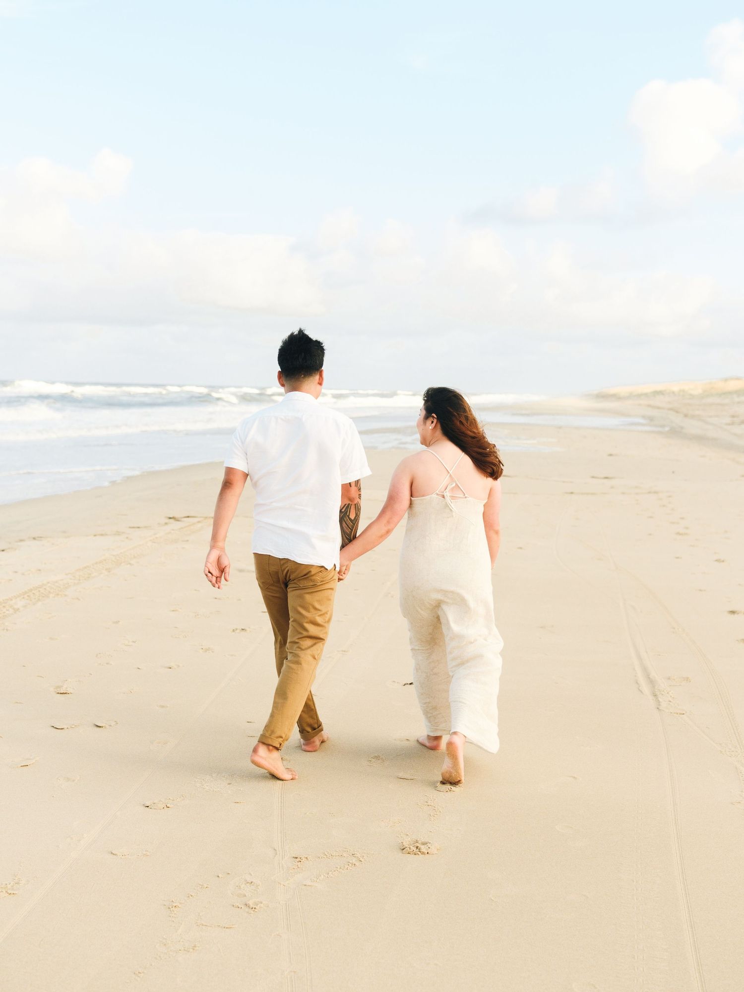 Couple in light colored outfits walks hand in hand along a serene sandy beach during golden hour.