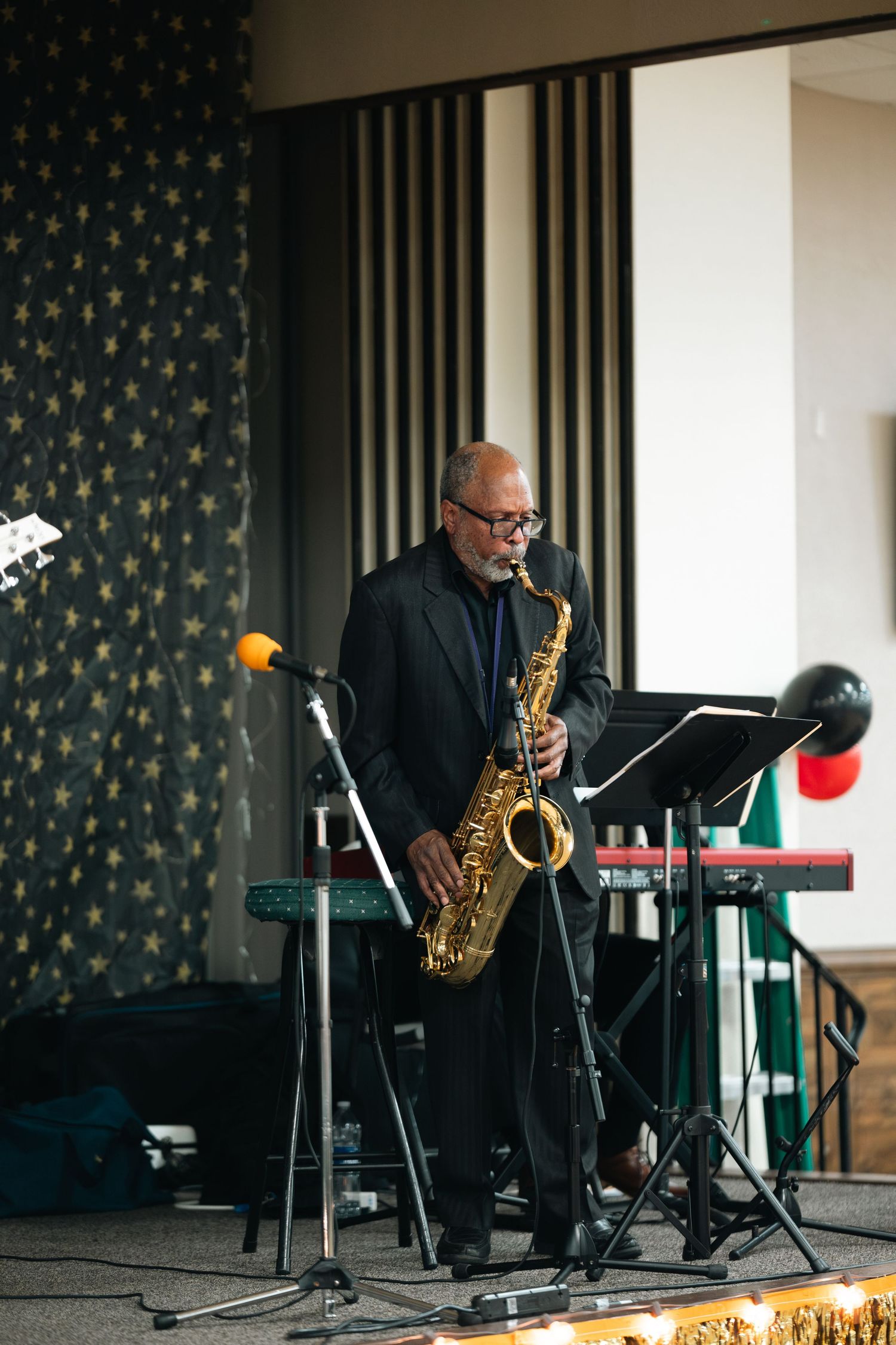 Musician performs on saxophone at an event with music stands and keyboard equipment on stage against patterned wallpaper.