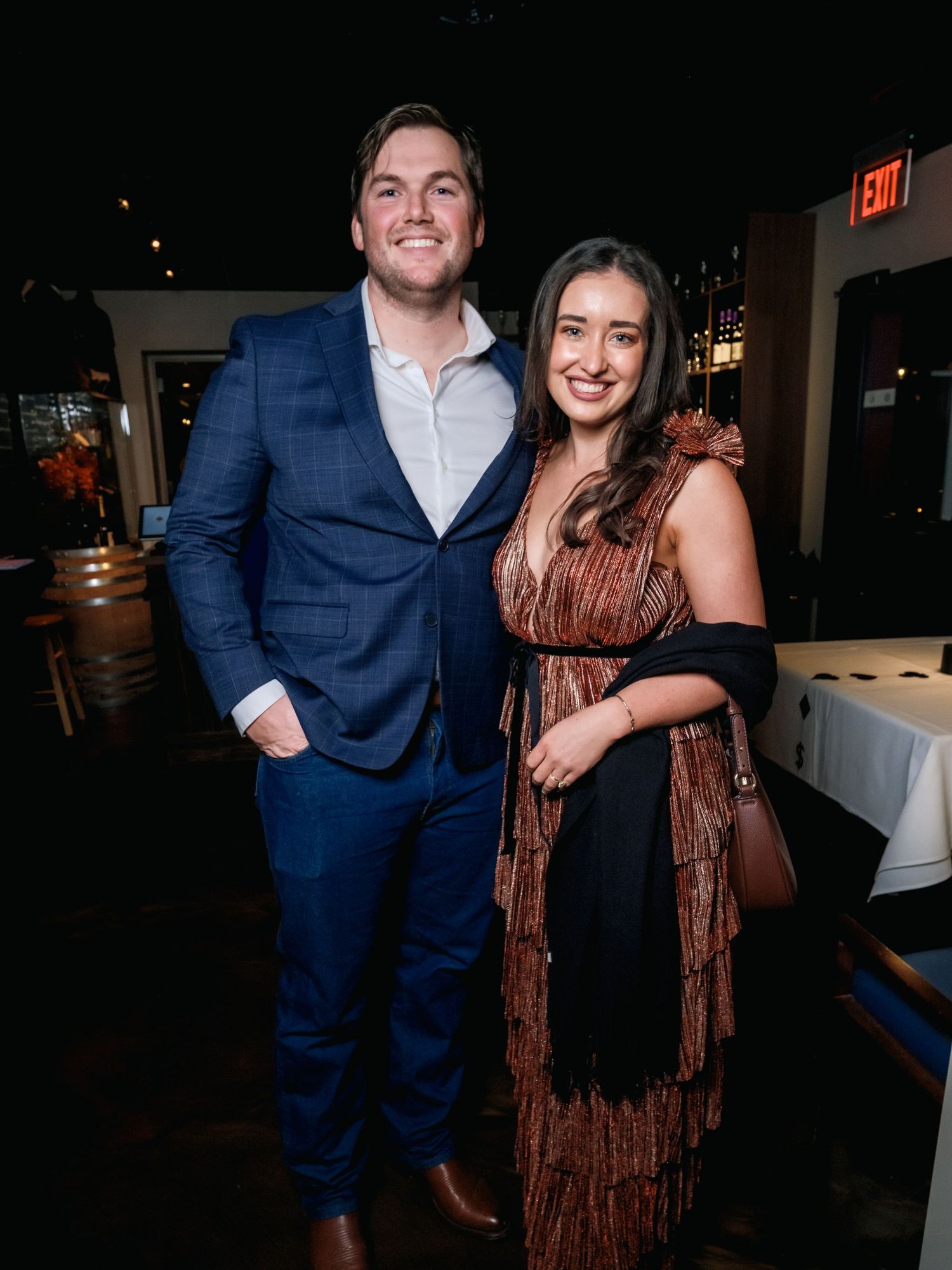 A couple dressed in formal attire poses together at an evening event with a navy suit and brown dress.