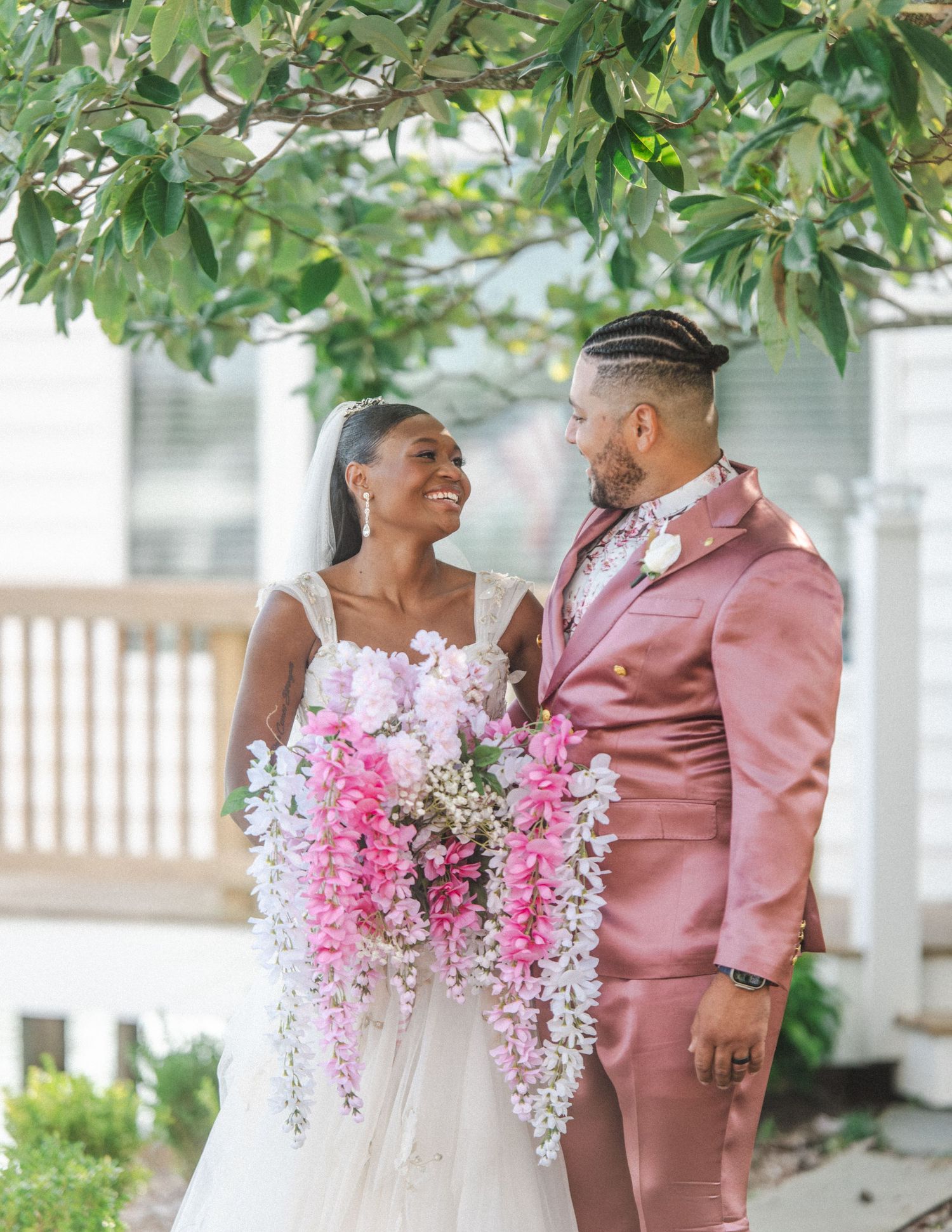 Newlywed couple sharing a joyful moment outdoors with cascading pink floral bouquet under green tree branches.