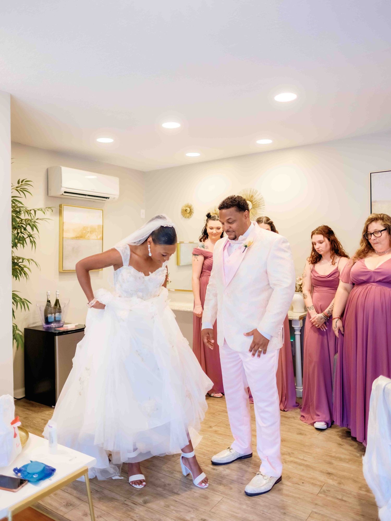 People gathered in a bridal suite with bridesmaids in purple dresses watching as the wedding couple share a moment.