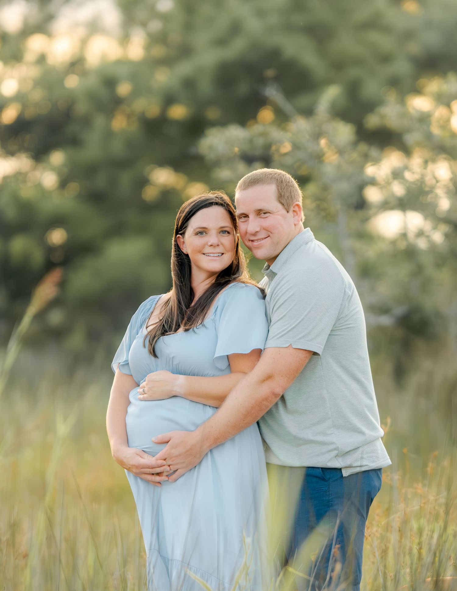 Couple poses together in a field at sunset during a maternity photo session in light blue outfits.
