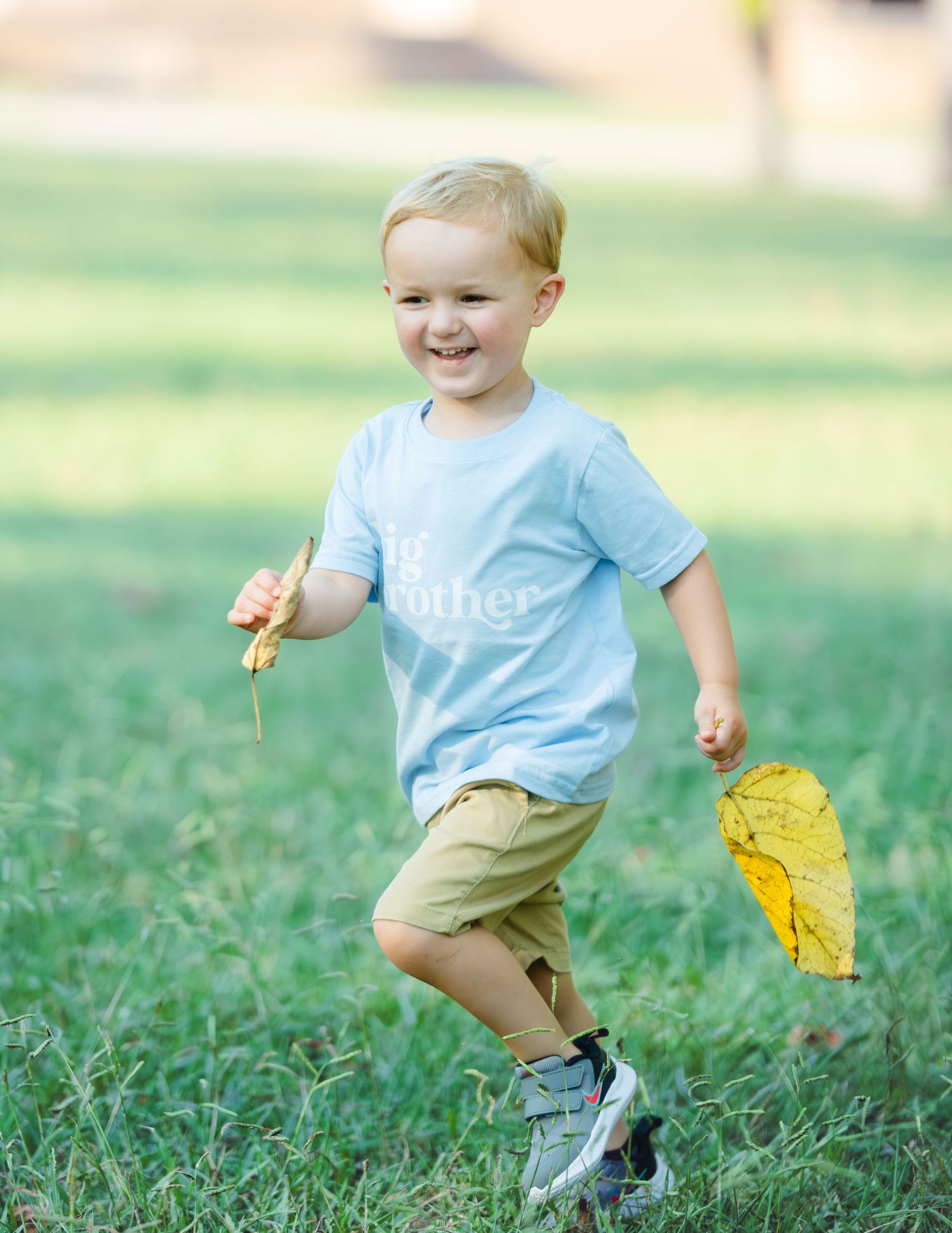 Young child wearing light blue shirt and khaki shorts runs on grass holding yellow autumn leaves.
