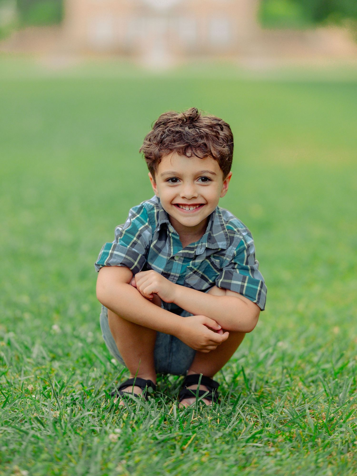 Child wearing plaid shirt crouches on green grass in series of candid outdoor portraits.