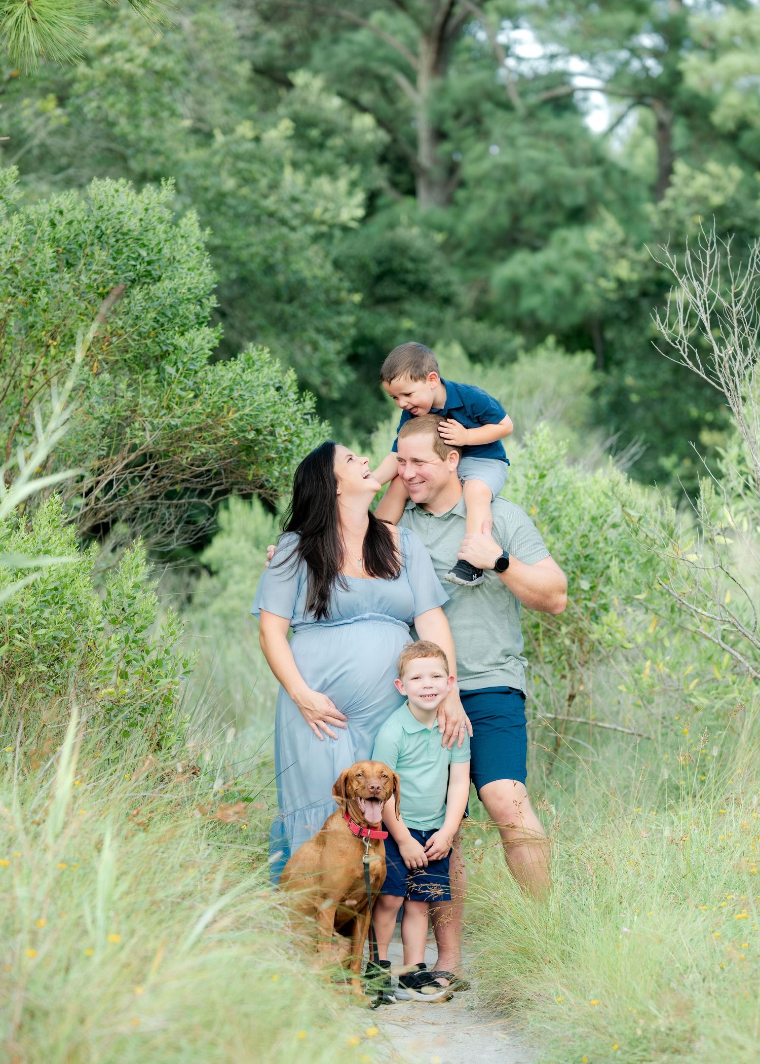 A family and their golden retriever pose together on a grassy trail surrounded by lush green foliage in a natural outdoor setting.