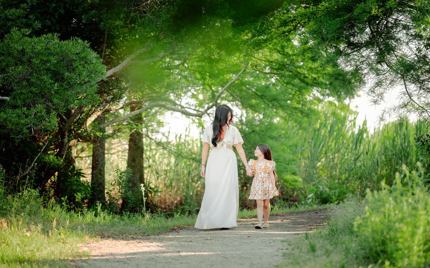 Two people walk down a sunlit garden path surrounded by lush green trees and wildflowers on a summer evening.