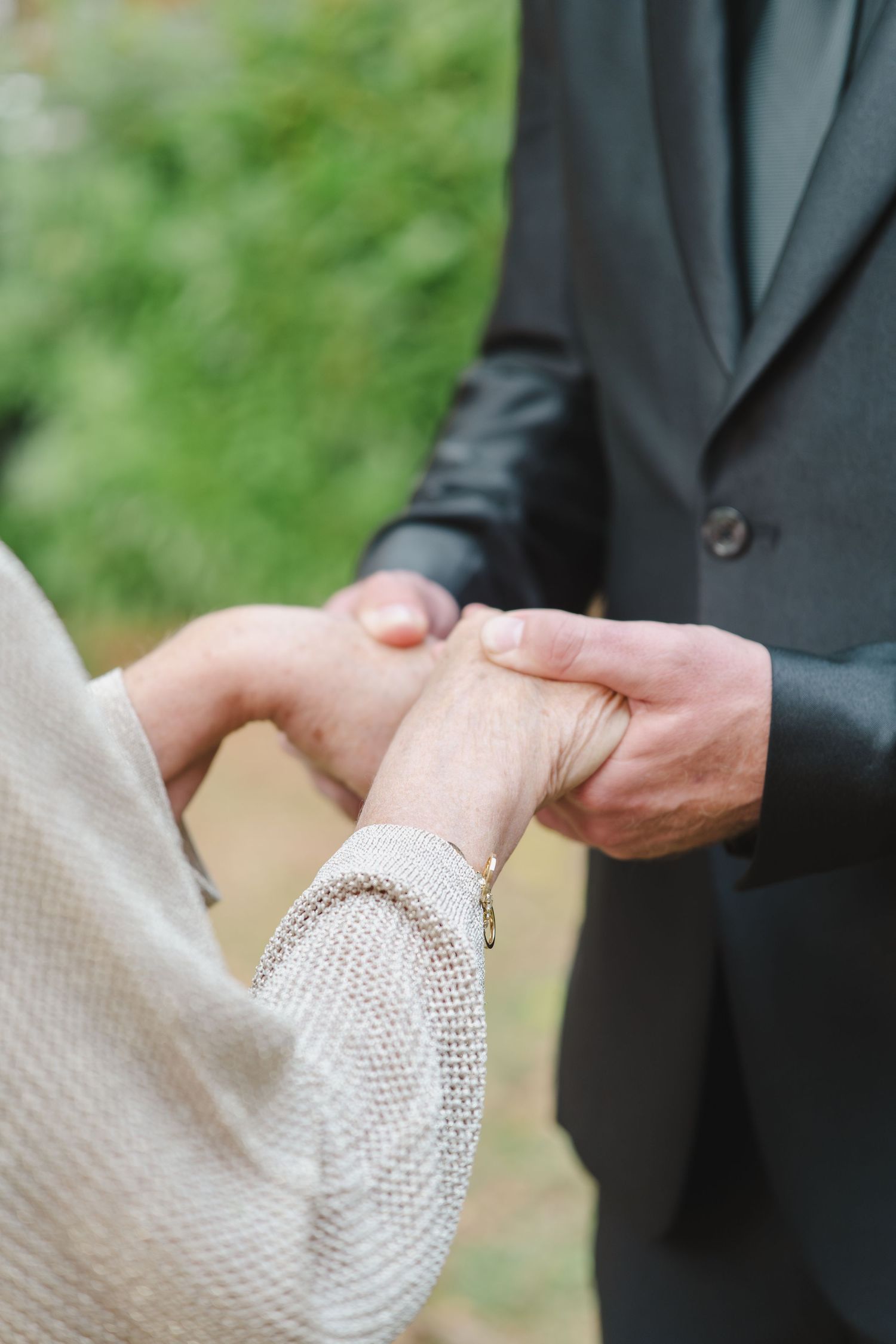 Close-up shot of hands exchanging wedding rings during an outdoor ceremony, with a dark suit and light sweater visible.