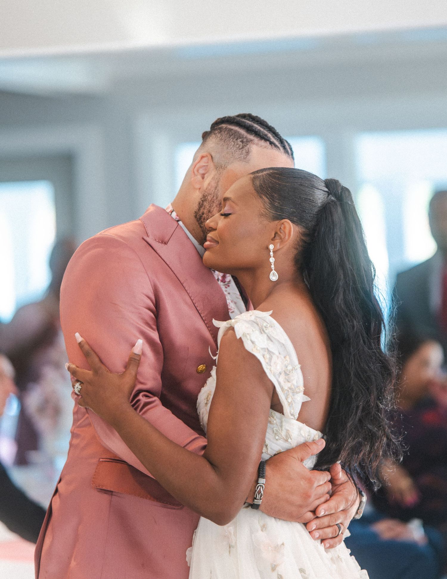 A couple shares an intimate moment during their first dance at a wedding reception, wearing pink suit and white dress.