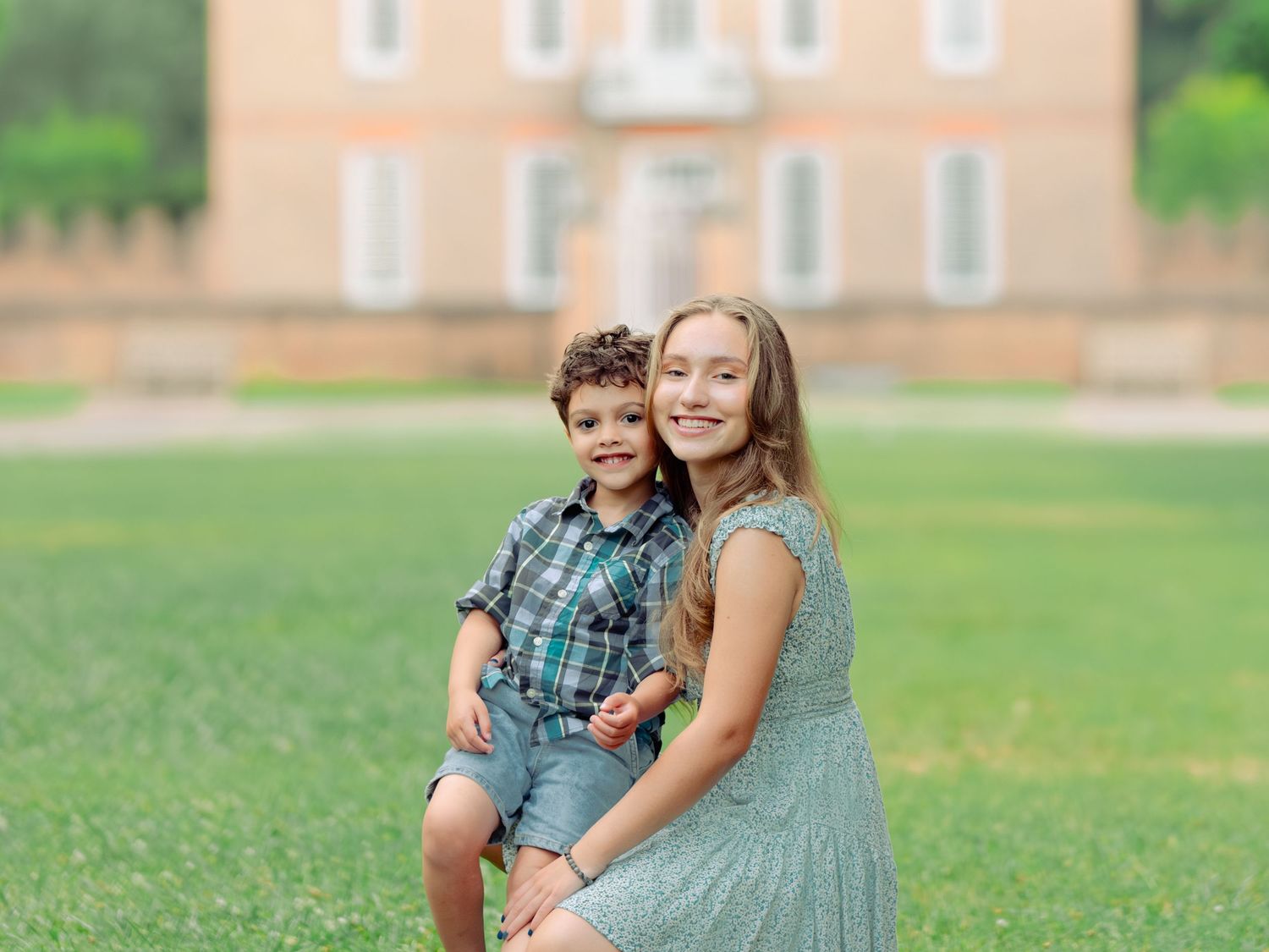 A parent and child sit together on a wooden bench in front of a peach colored building on a sunny summer day.