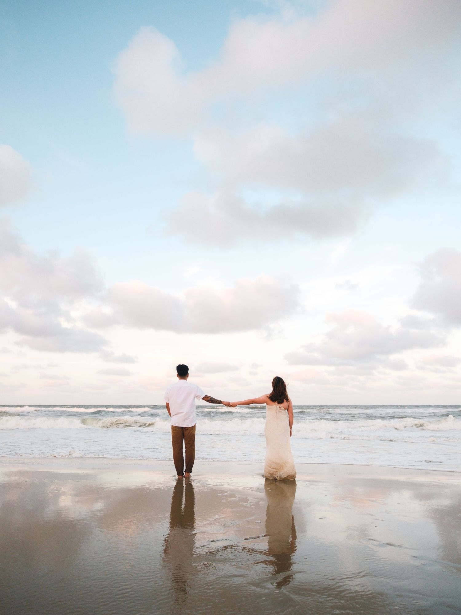 A couple stands together on a misty beach at sunset, walking along the shoreline with waves rolling in.