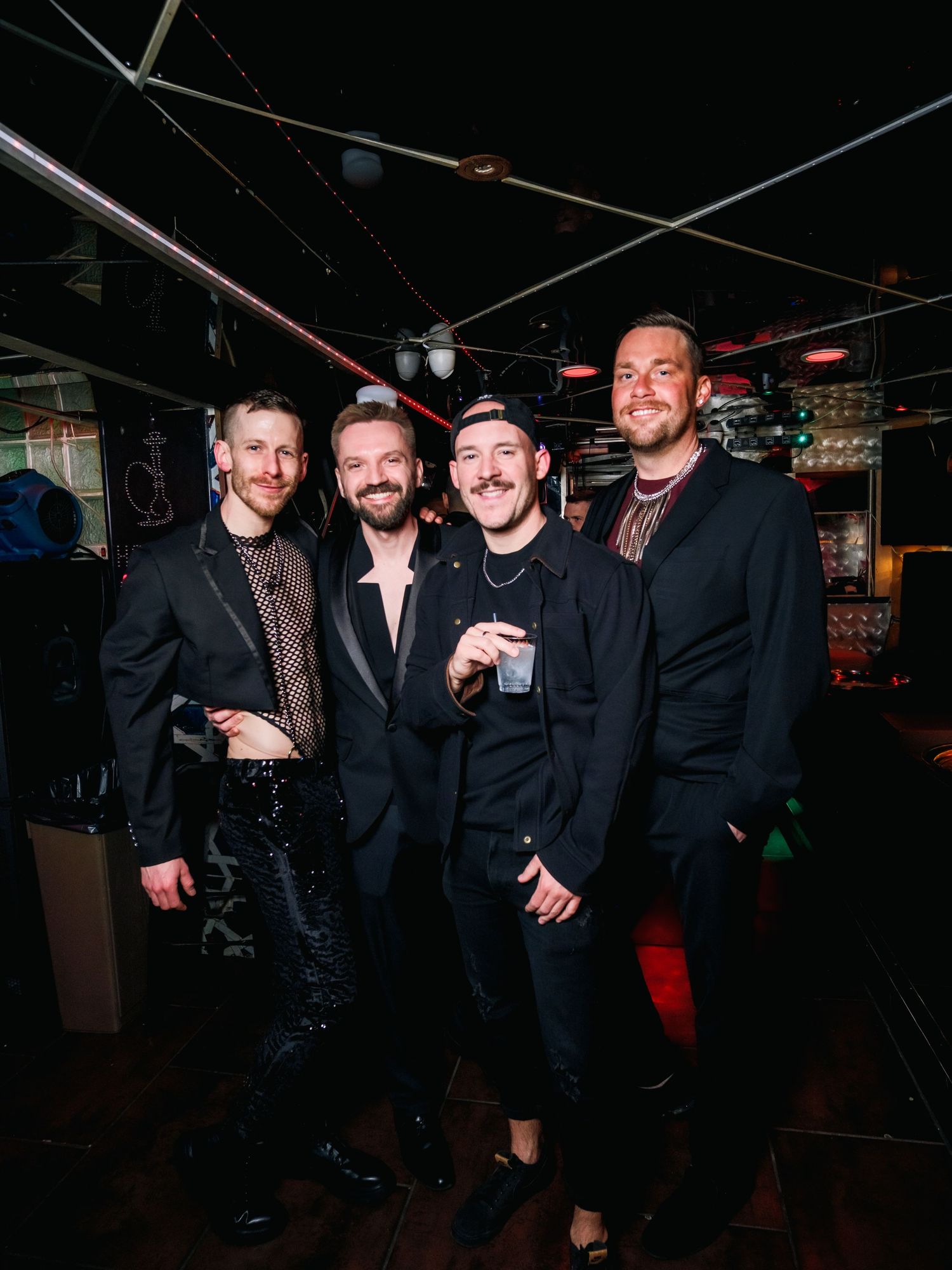 Four people in dark formal attire pose together at a dimly lit venue with exposed ceiling lighting.