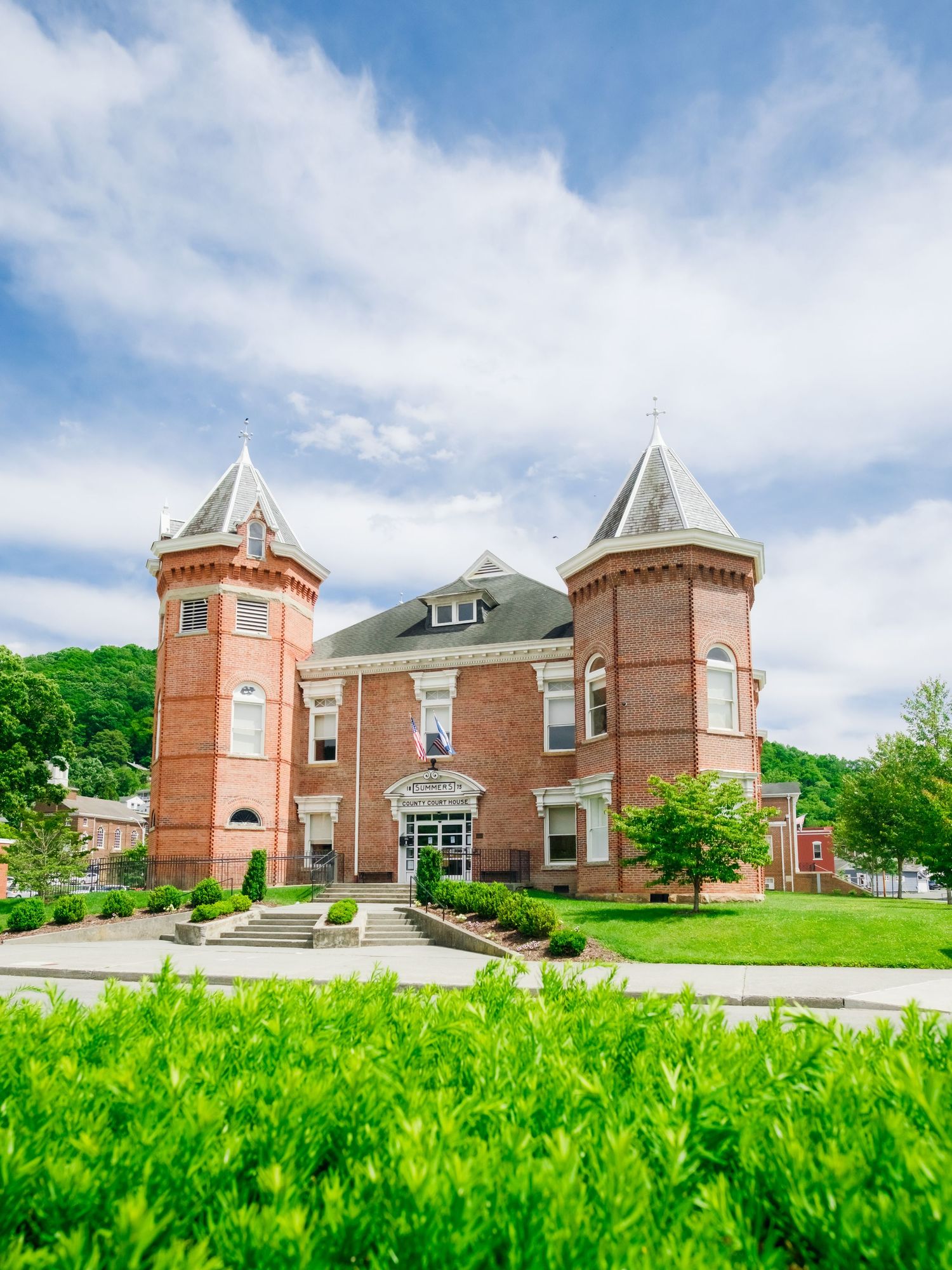 Historic red brick church building with turrets and expansive green lawn under a bright blue cloudy sky.