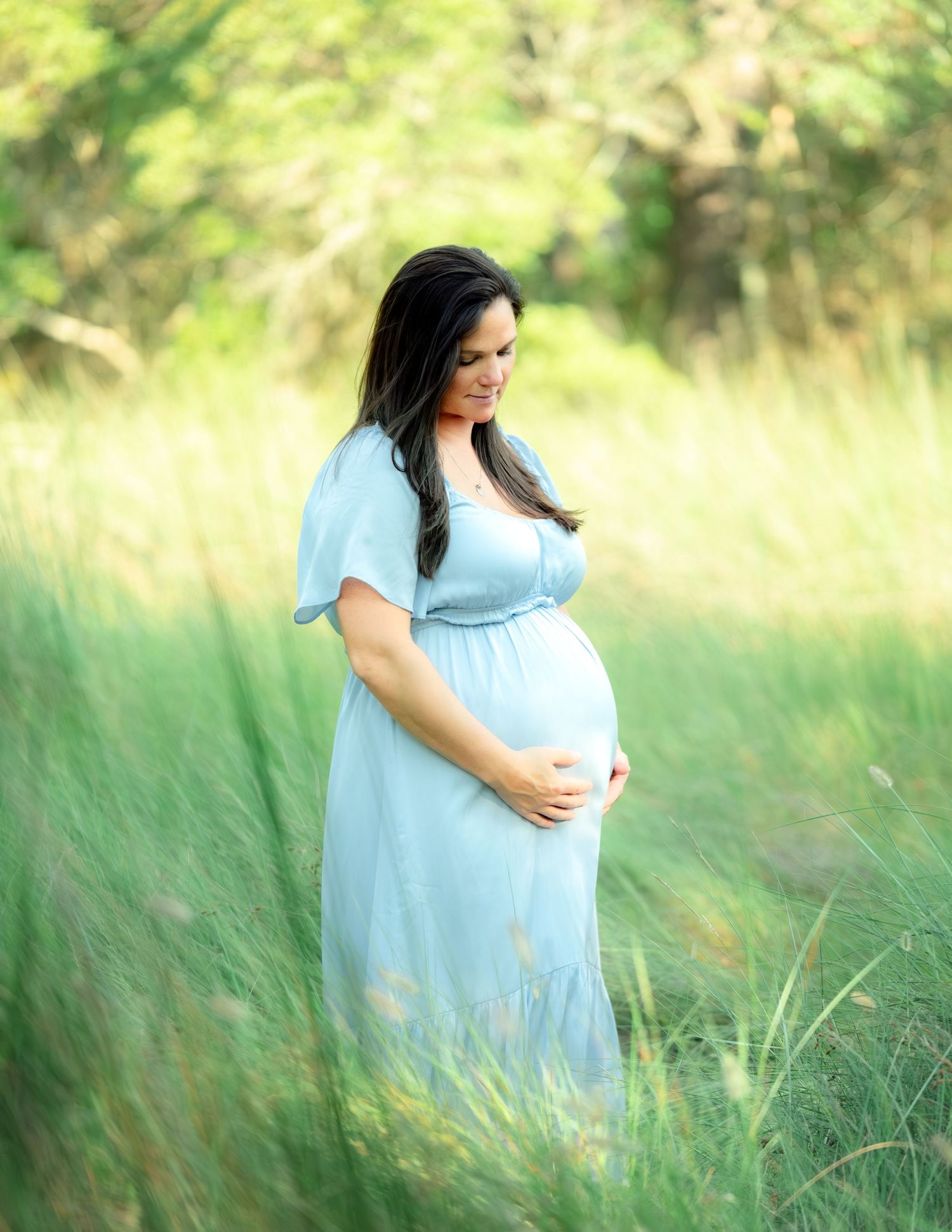 Expectant figure in a light blue flowing dress stands serenely among tall grasses in a sunlit meadow.