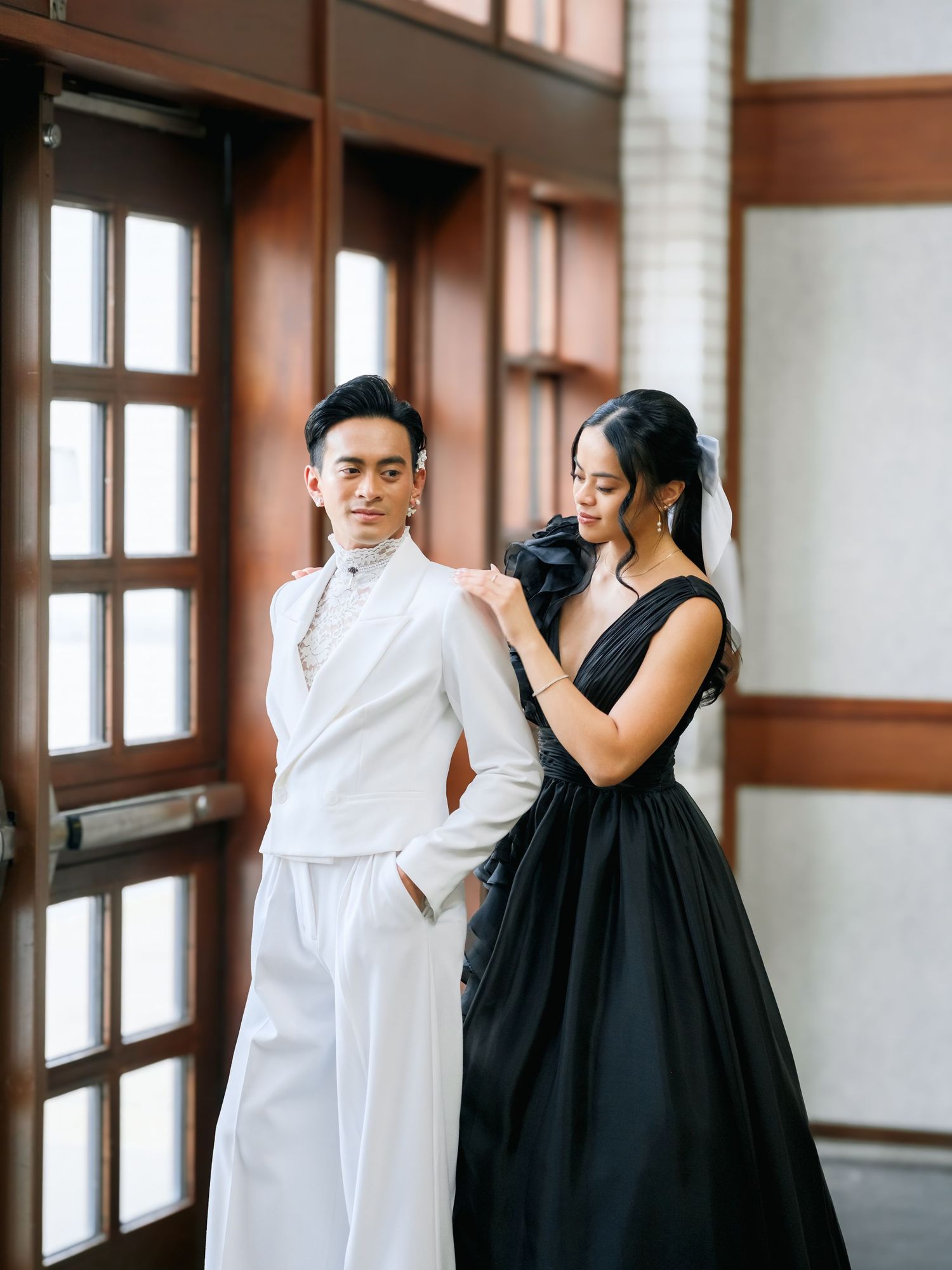 A couple in black and white formal attire dance together in an elegant windowed hallway.