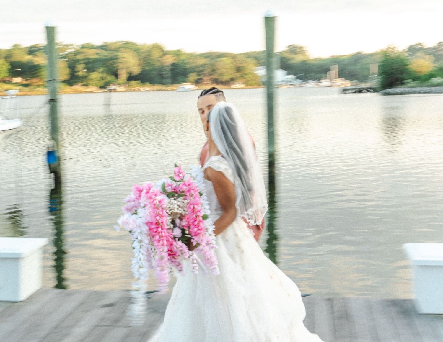 A bride in a flowing white gown holds a vibrant pink bouquet while standing on a dock overlooking the water.
