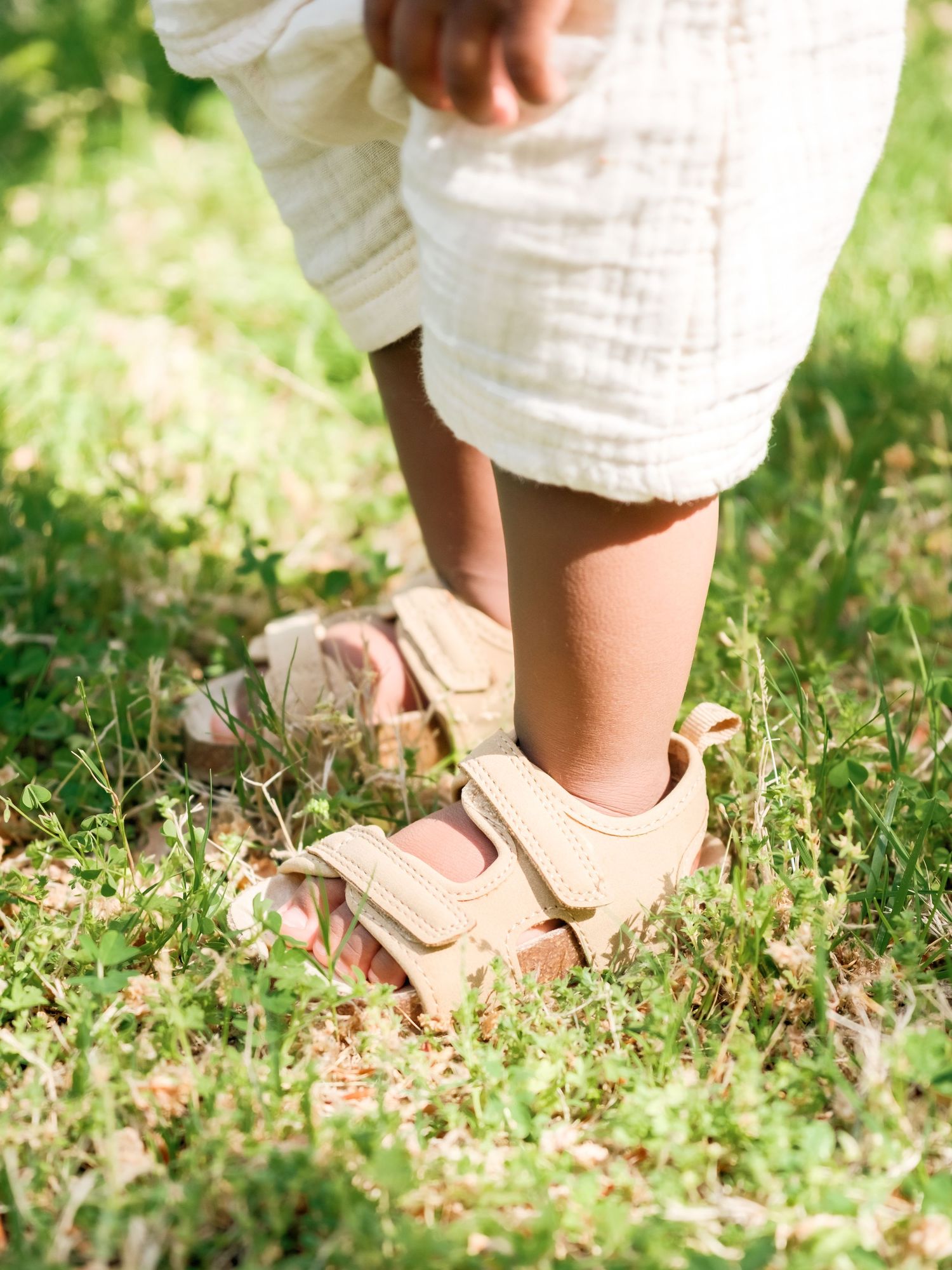 Little feet in beige velcro sandals standing on grass wearing white linen clothing on a sunny day.