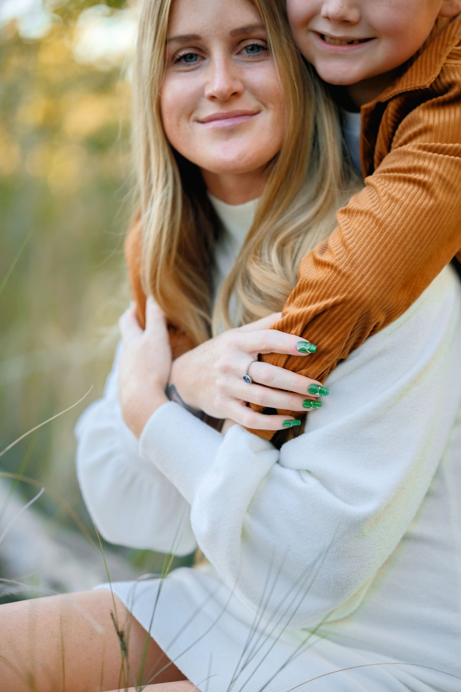 A couple in white and brown sweaters embraces outdoors in soft autumn lighting.