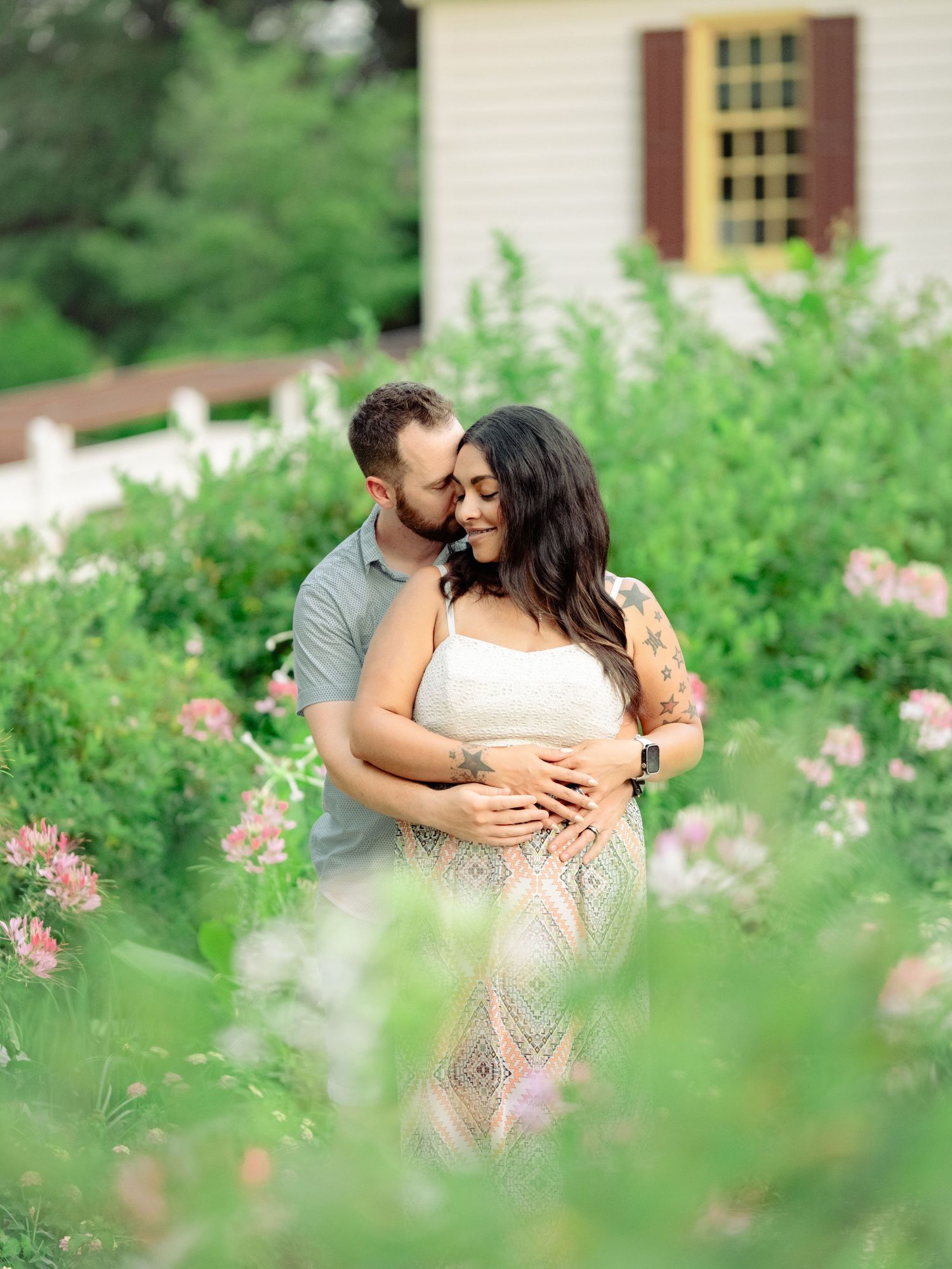 A romantic couple embraces in a lush garden with pink flowers and a white house with shutters in the background.