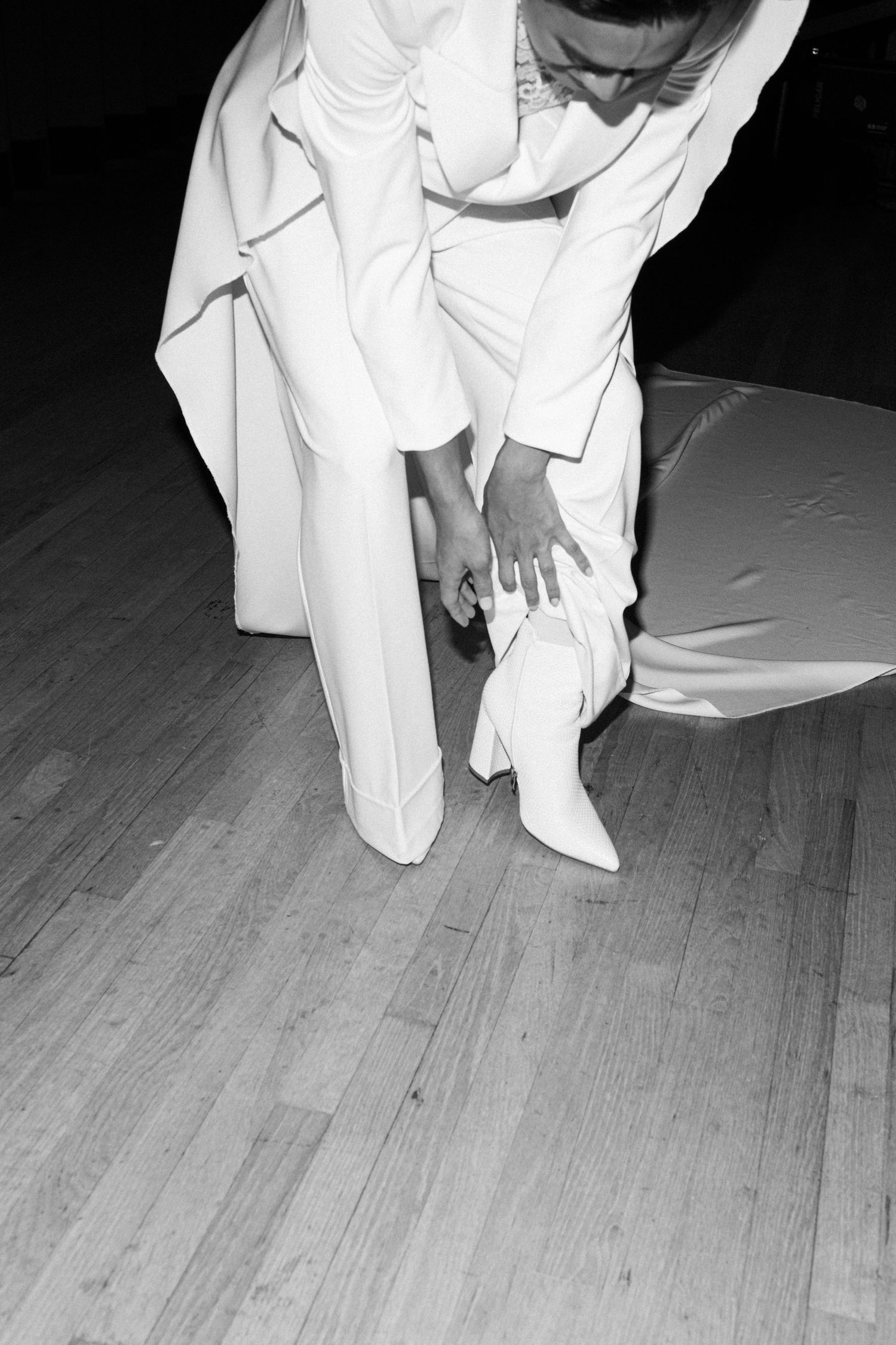A person in white clothing bends down to adjust their white boots on a wooden floor during a formal occasion.