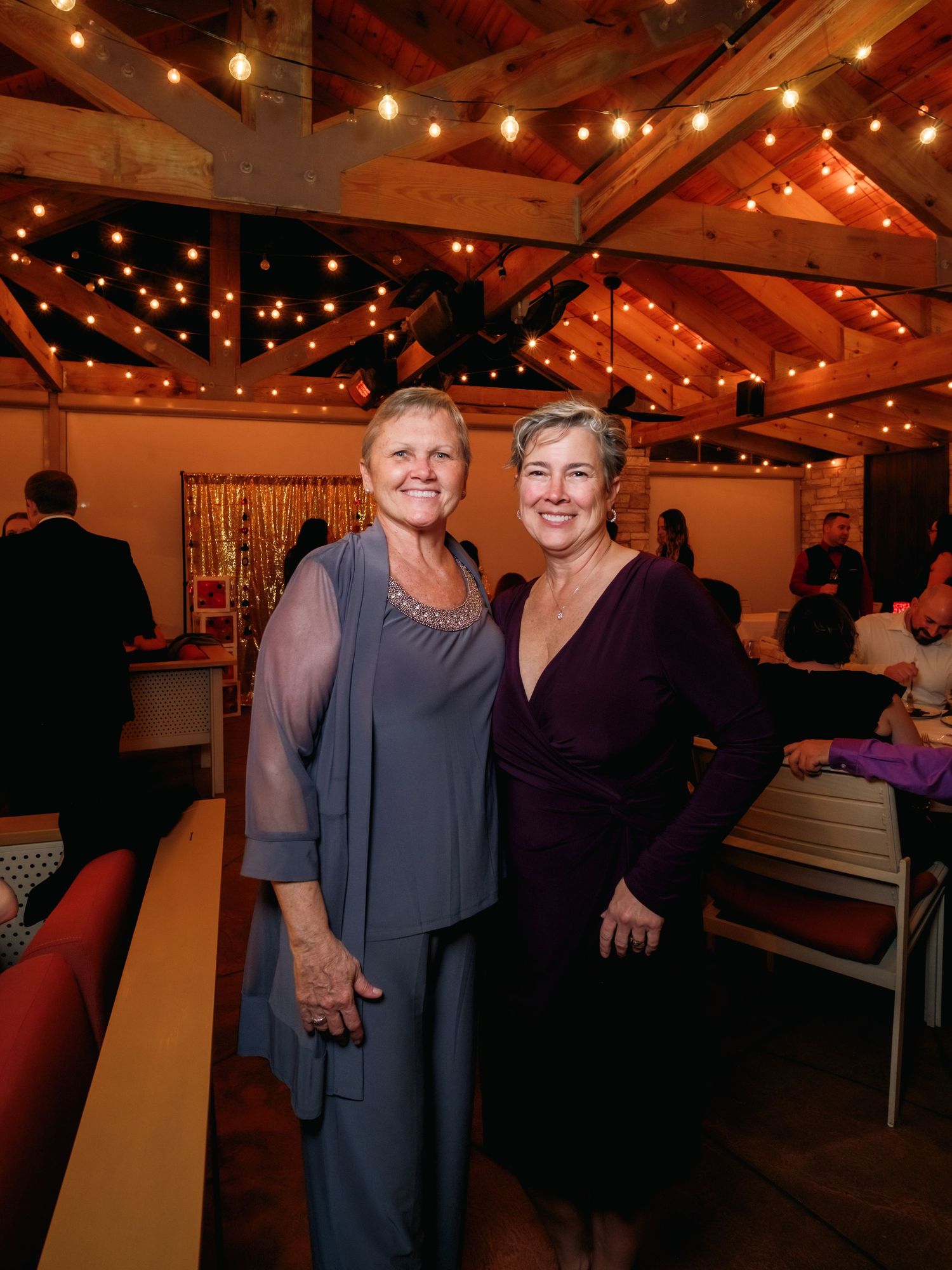 Two people pose together at a celebration under rustic wooden beams with string lights.
