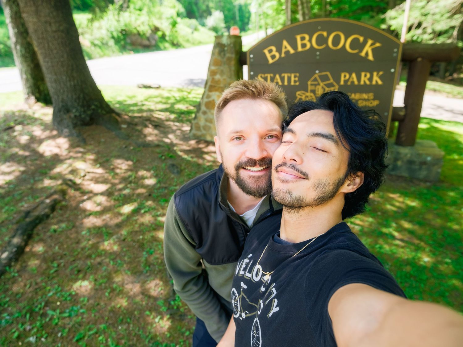 Two people take a smiling selfie in front of a wooden Babcock State Park entrance sign on a sunny day.