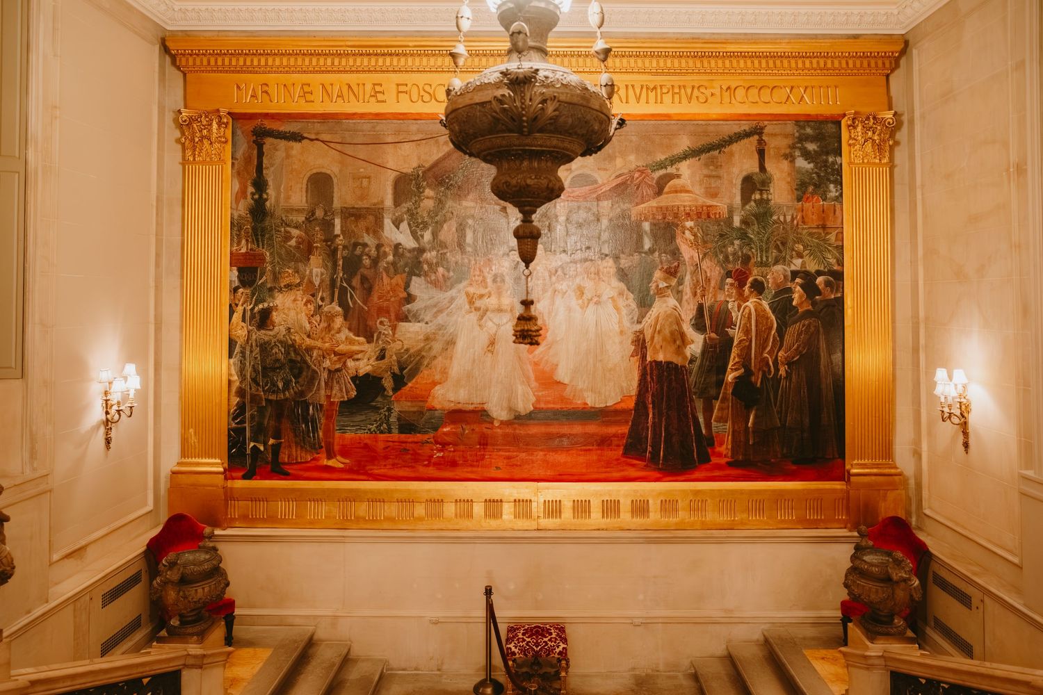A grand hallway with ornate gold-framed paintings and wall sconces features elegant classical decor and marble floors.