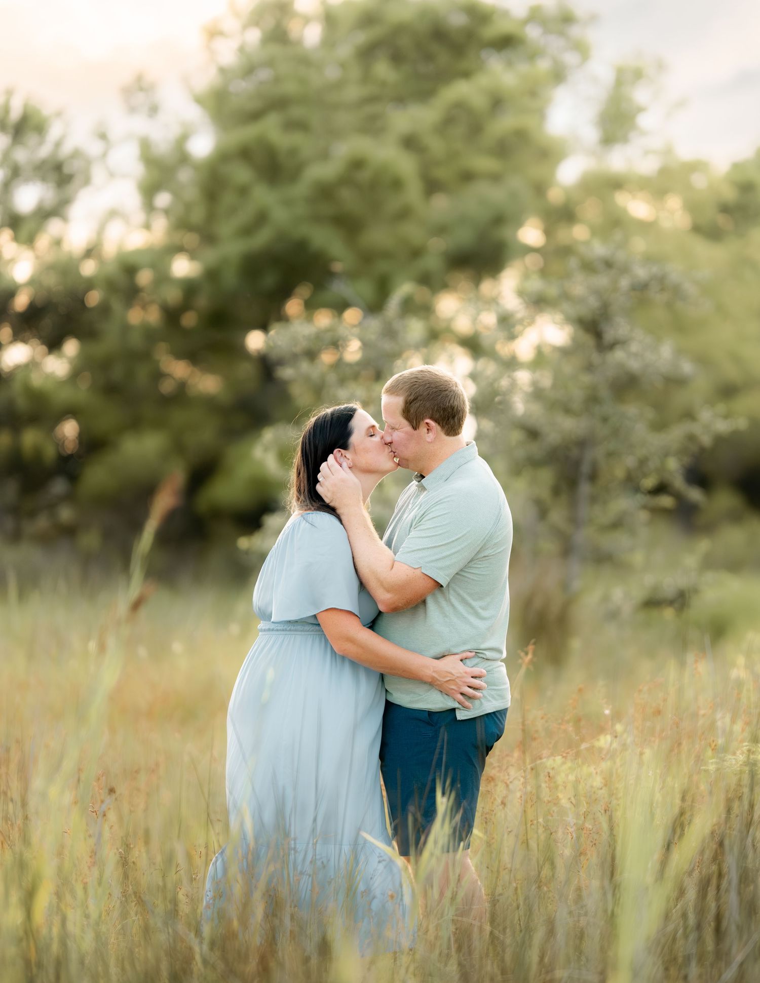 A couple in blue and mint outfits embrace tenderly in a golden field during sunset time.
