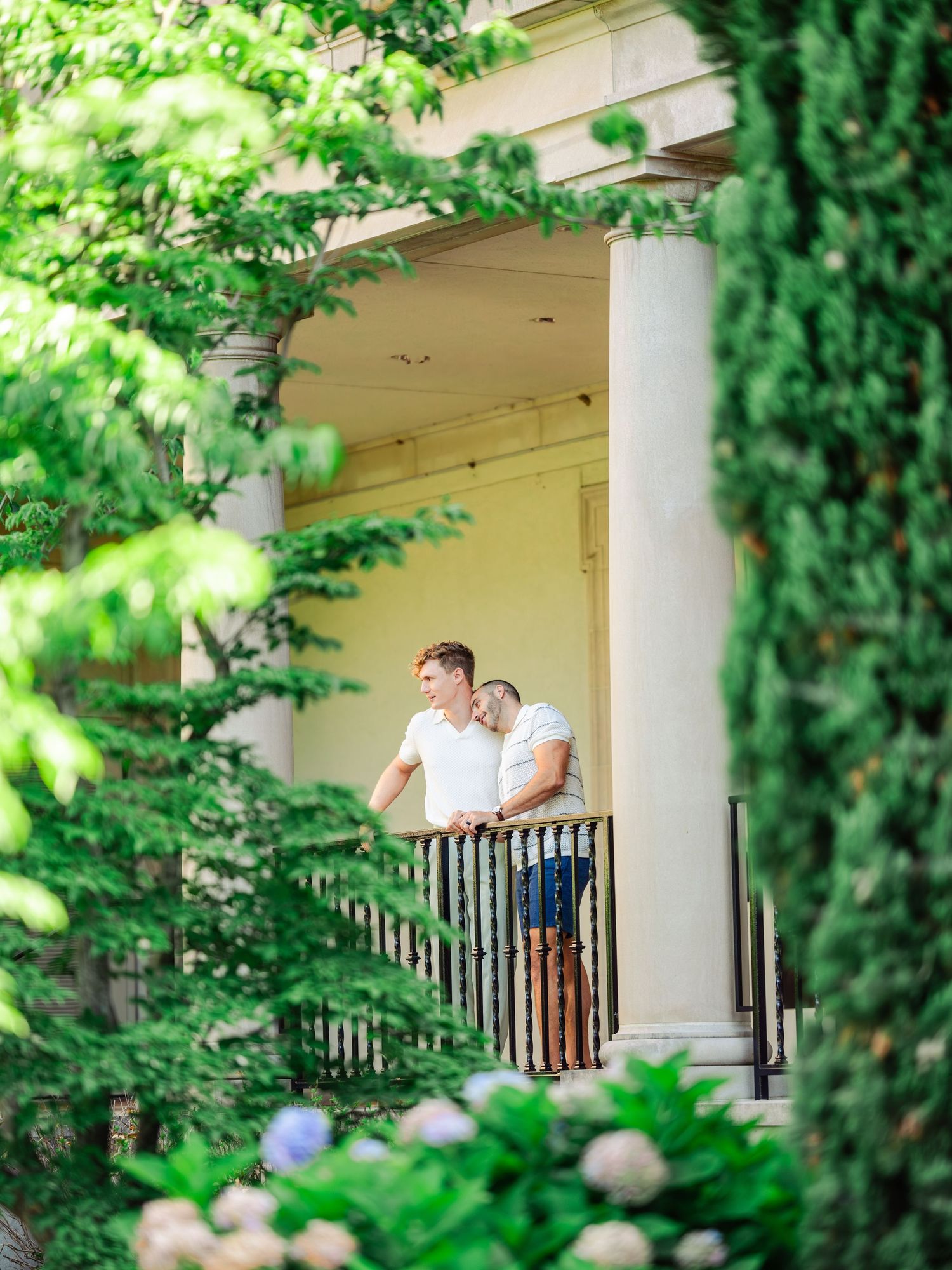 A couple stands together on a white-columned porch surrounded by lush green foliage and garden landscaping.