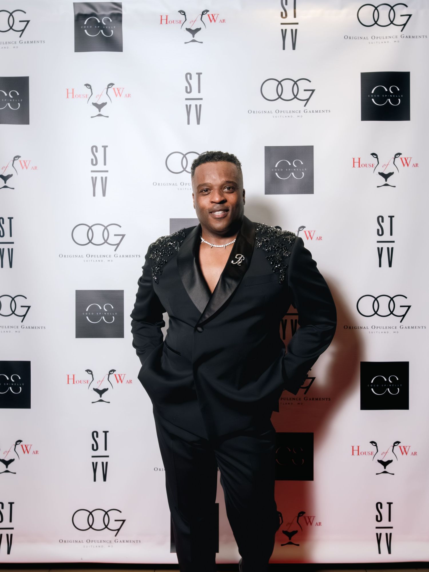 A person poses in a black embellished suit against a step-and-repeat backdrop with various logos and branding.