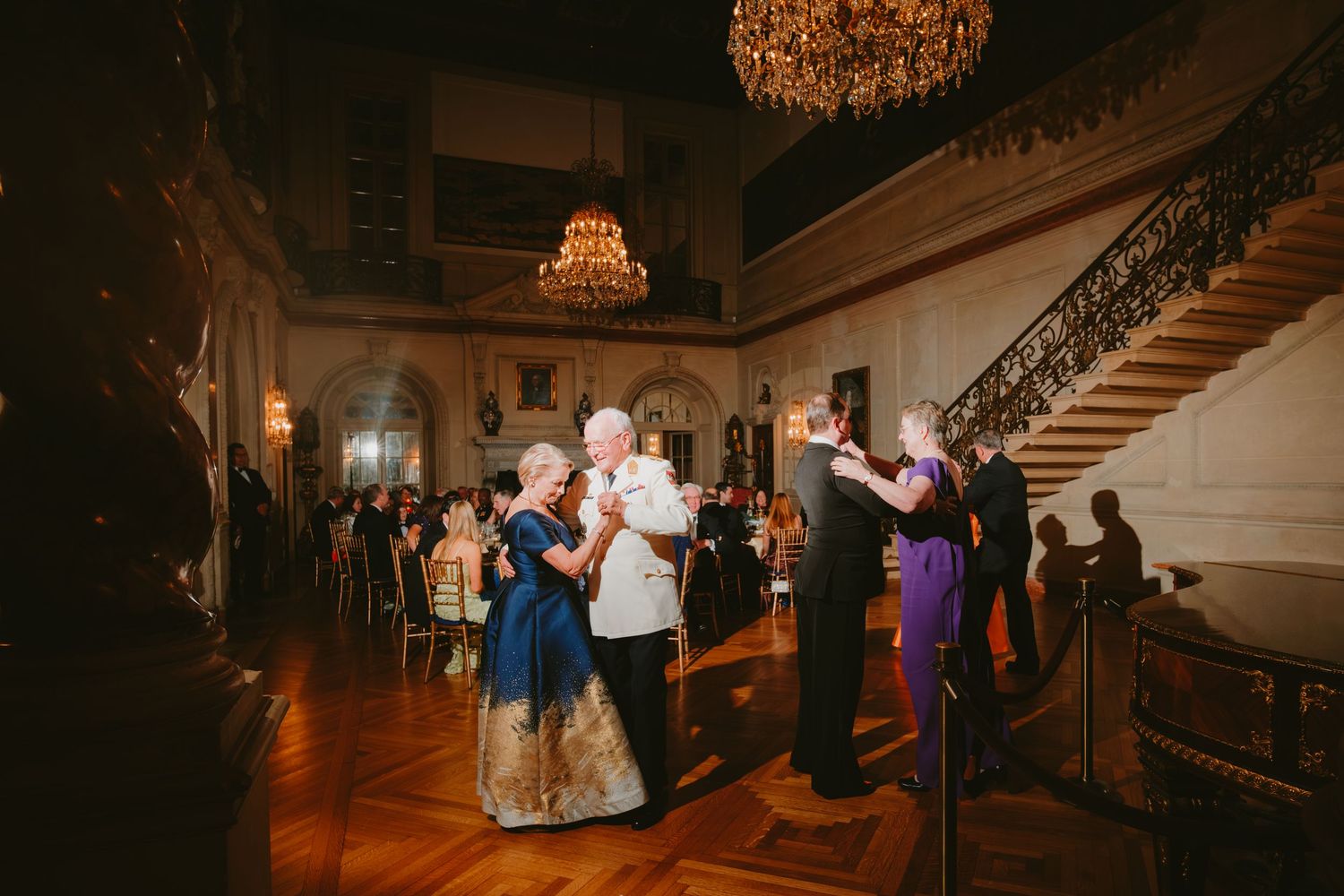 Couples dancing together in an elegant ballroom with ornate chandeliers and a grand staircase in warm lighting.