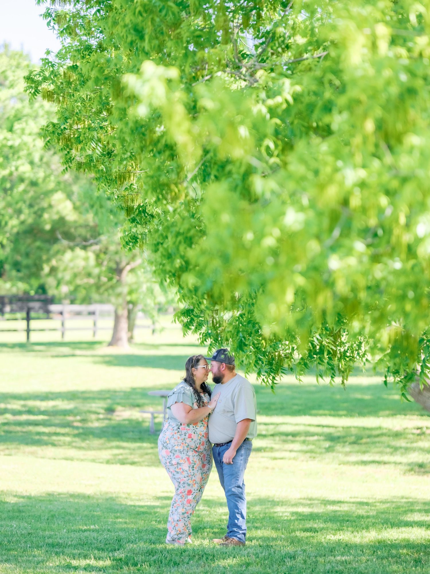 A couple shares a tender moment under green trees in a peaceful park setting on a sunny day.