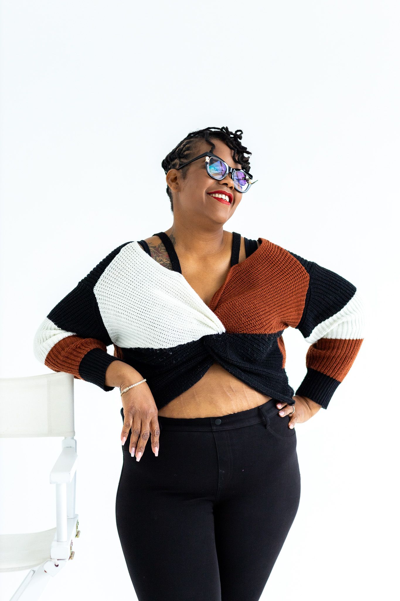 Fashionable person in sunglasses wearing a colorblock sweater in brown, black and white with black pants against white wall.