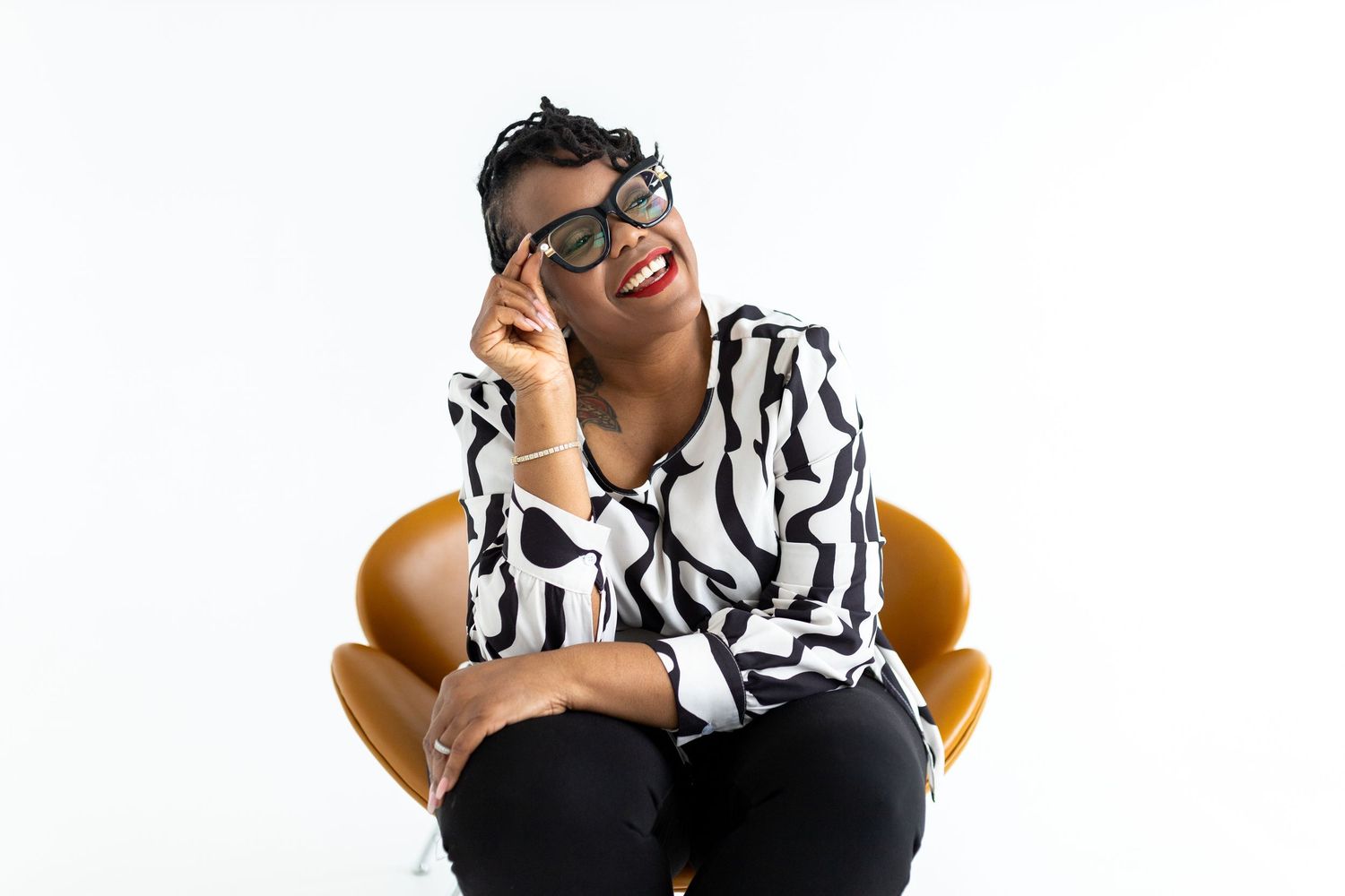 Person in black and white patterned shirt sits in orange chair while laughing and adjusting glasses.