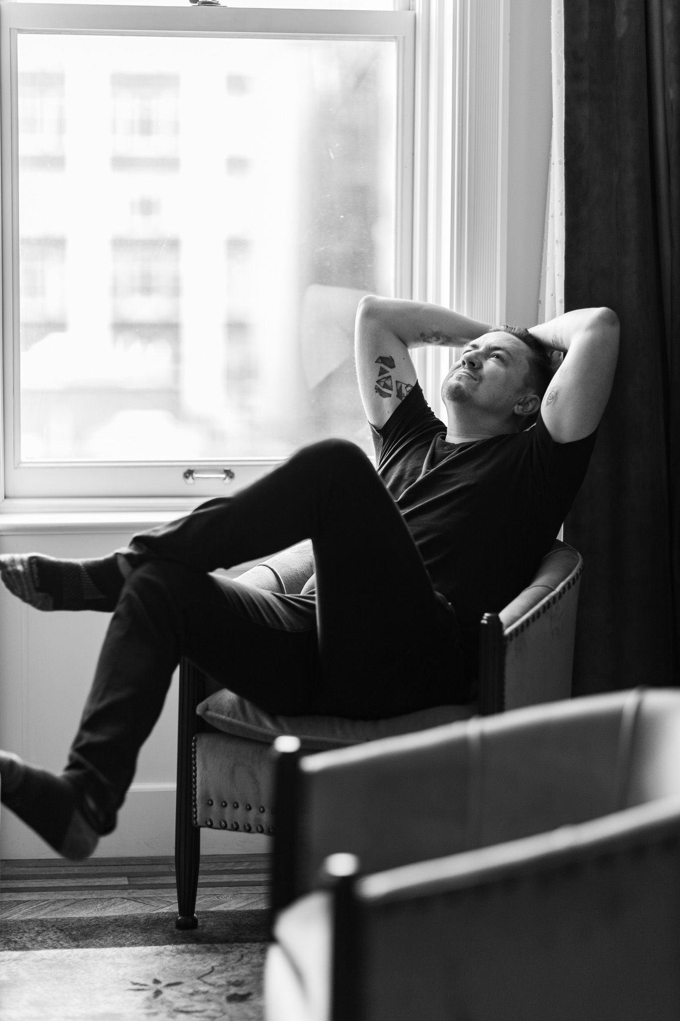 Black and white photo showing person relaxing in armchair by window in contemplative pose.