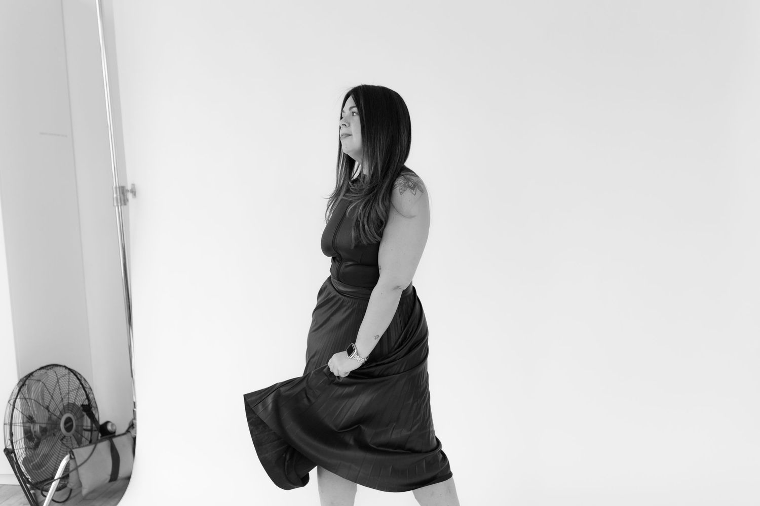 A black and white photo shows someone posing in a dark dress next to an electric fan.