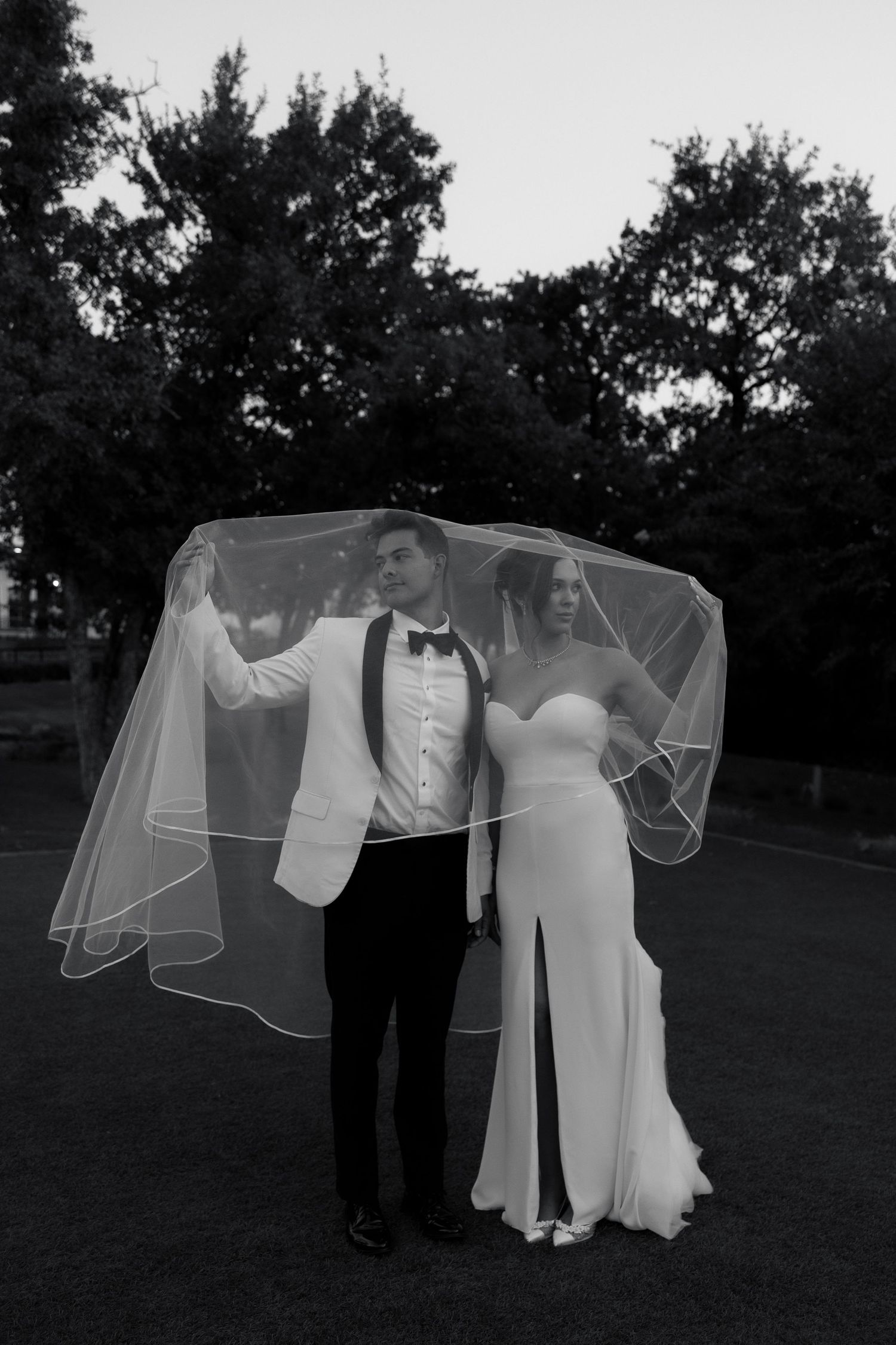 A series of black and white wedding photos showing a couple playing with a flowing veil in an outdoor garden setting.