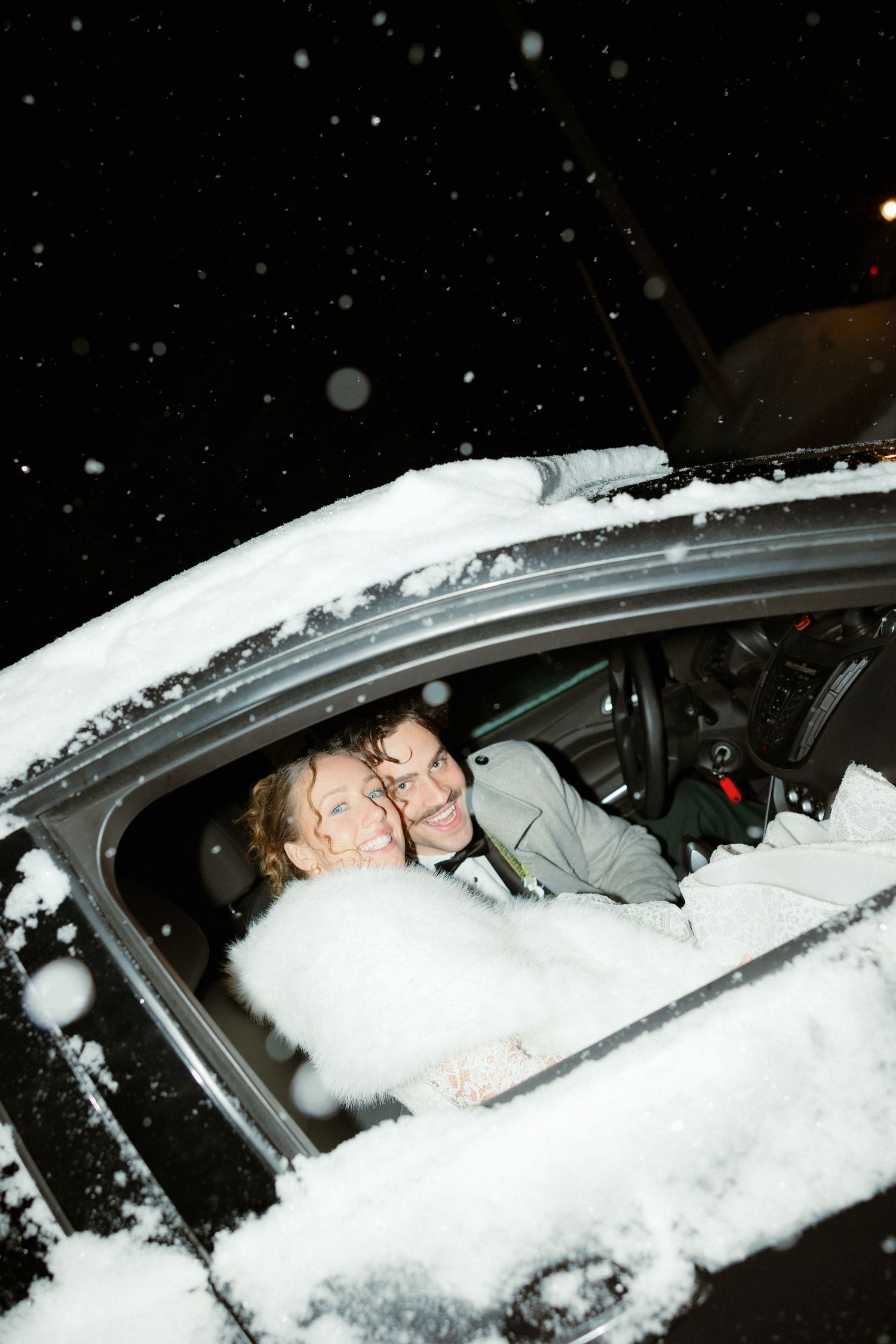 A couple sits in a snow-covered car at night with snow falling around them.