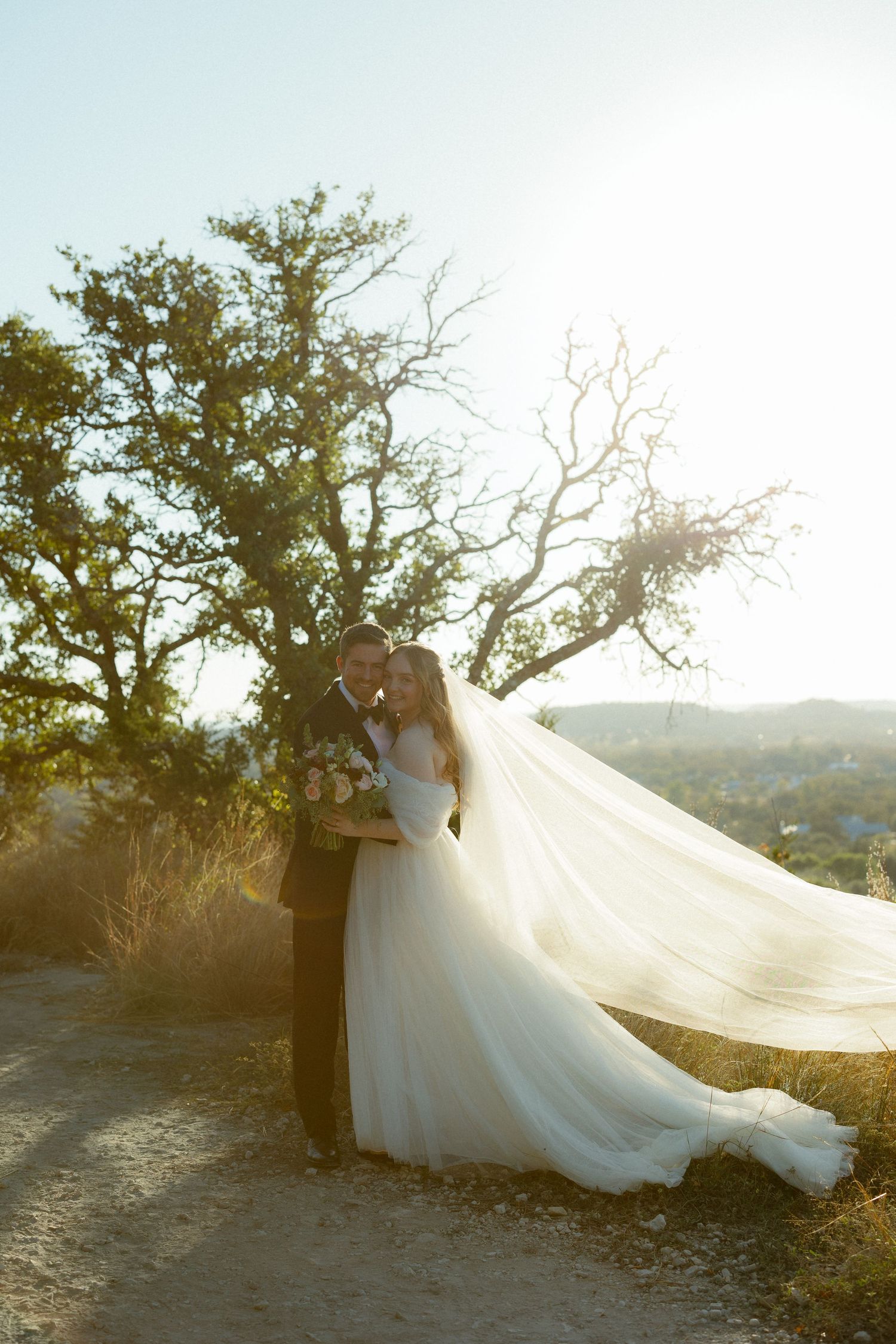 A couple in wedding attire embrace on a hilltop path at sunset as the bride's long veil flows dramatically in the breeze.