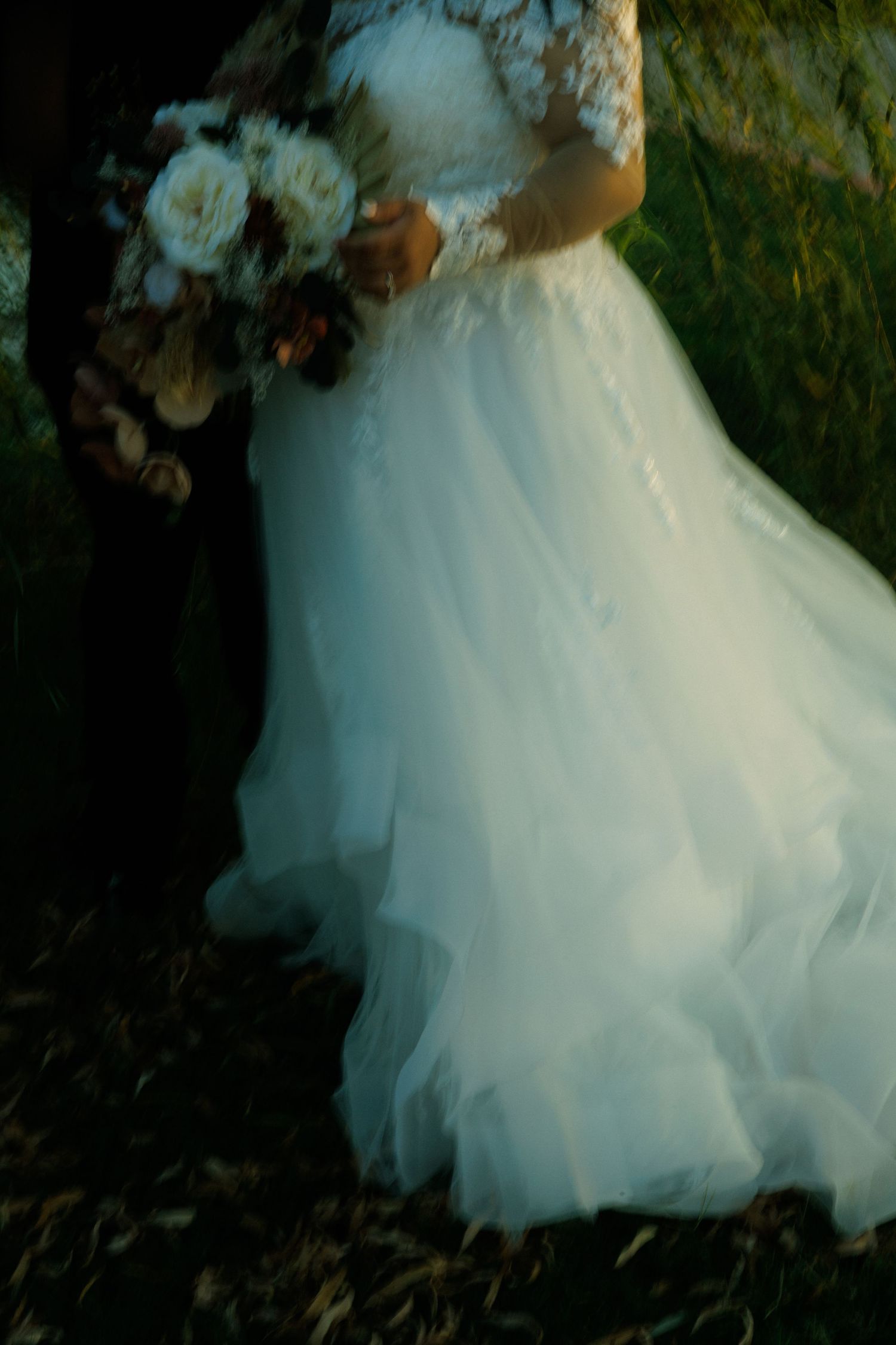 A flowing white wedding dress with a tulle skirt and floral bouquet captured in dreamy light against foliage.