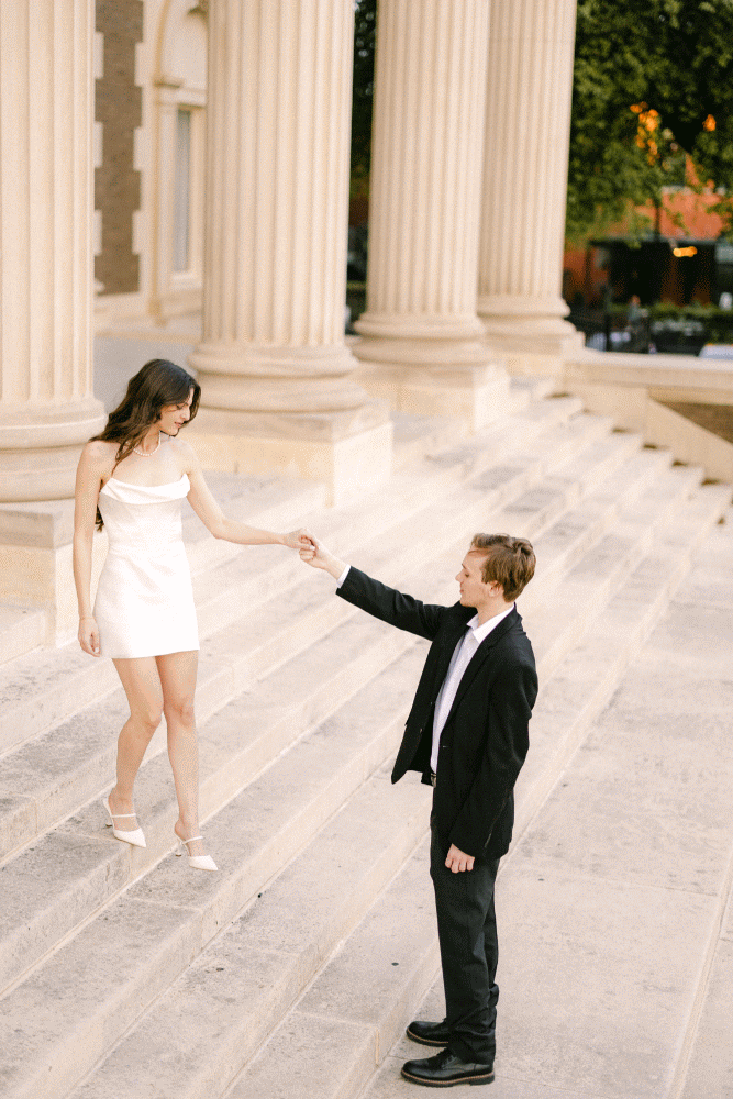 Romantic couple holding hands on marble steps with classical columns in background.